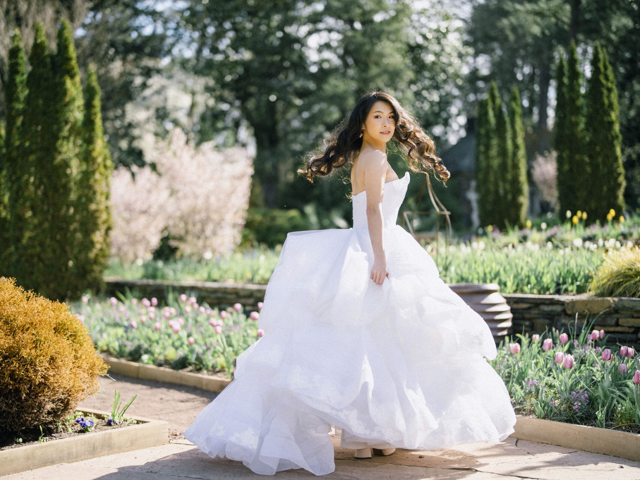 During her bridal session, a woman in a flowing white wedding dress twirls joyfully in a garden. The background is filled with blooming flowers, vibrant greenery, and tall trees under a clear sky.