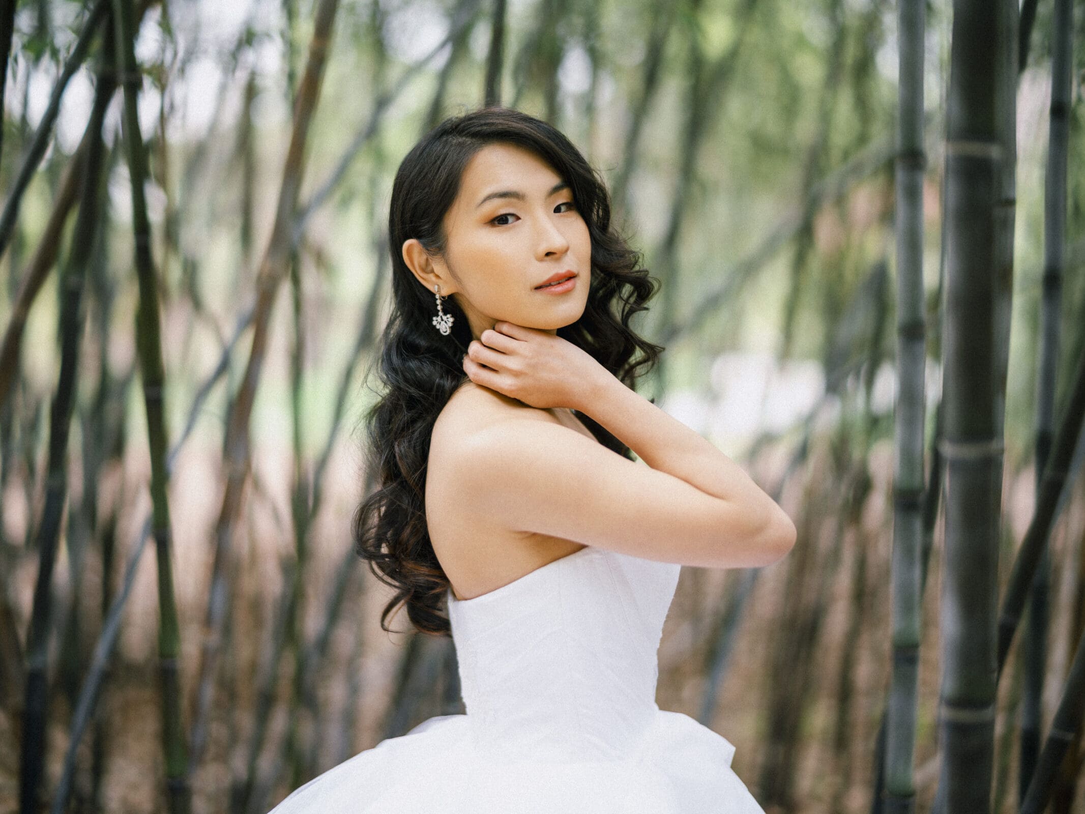 Amidst the serene bamboo forest, a woman in a strapless white dress poses gracefully, embodying the essence of bridal sessions. Her long, wavy hair cascades as she touches her neck with one hand, casting a soft glance over her shoulder.