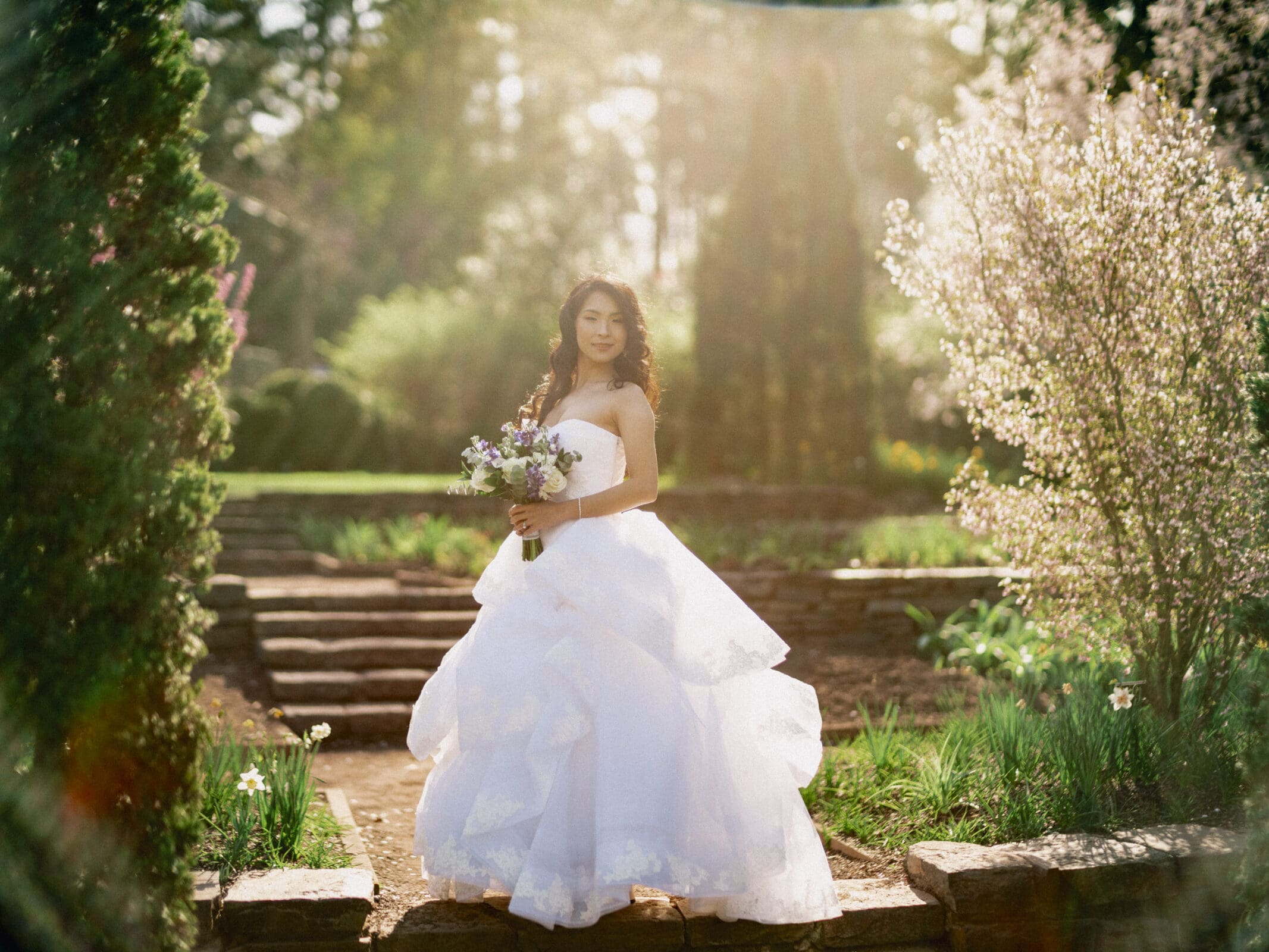 In a dreamy bridal session, a bride in a strapless white wedding dress holds a bouquet while standing on a stone path, surrounded by lush greenery and blooming trees. Sunlight filters through the foliage, casting an enchanting glow over the scene.