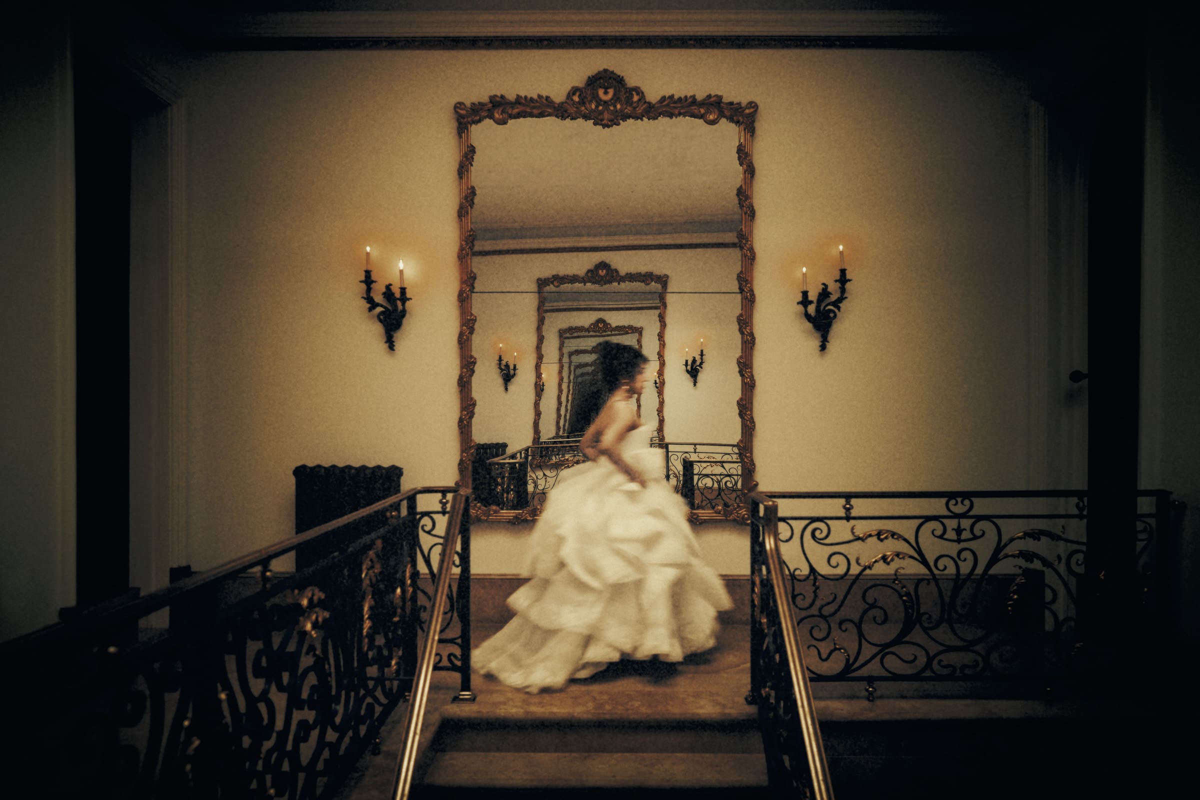 A person in a white, ruffled dress walks down a dimly lit hallway, the setting reminiscent of elegant bridal sessions. The ornate mirrors create reflections that repeat into the distance, while the vintage ambiance is accentuated by decorative wall sconces.