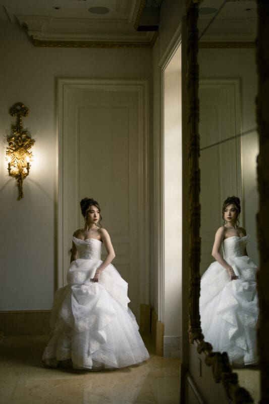 A bride, during her bridal session, stands near a mirrored wall in an elegant room, wearing a white, strapless wedding gown with a voluminous skirt. Soft lighting casts a warm glow, highlighting her reflection in the mirror and the ornate wall sconce beside her.