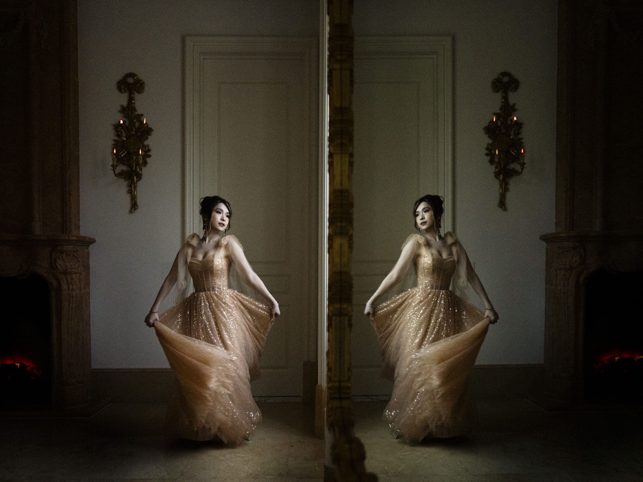 A woman in an elegant, sparkling gown poses gracefully in a dimly lit room, evoking the charm of bridal sessions. She stands near an ornate mirror, creating a vivid reflection. The room's warm ambiance is enhanced by two decorative sconces on the walls.