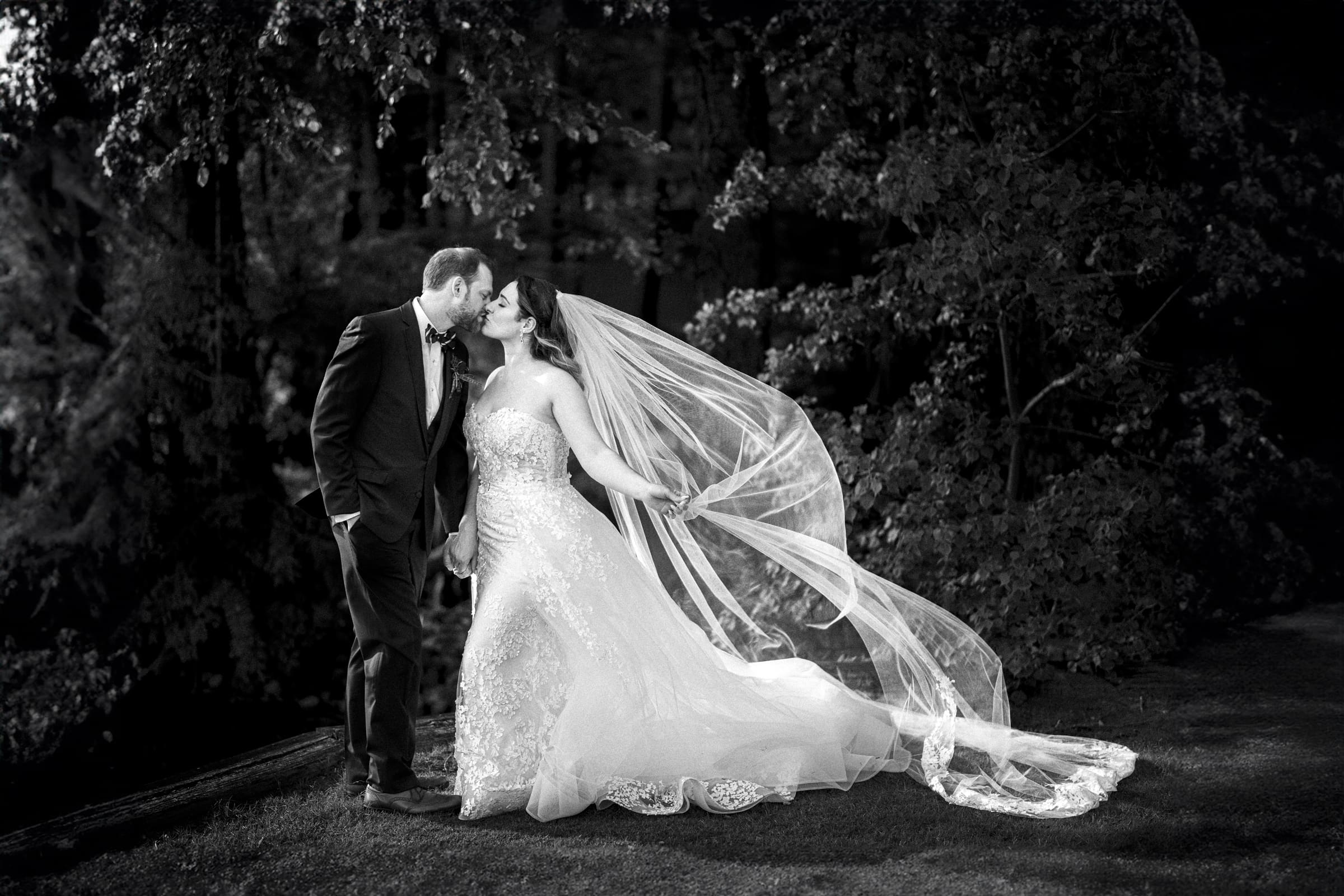 A black and white image of a bride and groom at a river landing. The groom, in a suit, gently kisses the bride on the forehead. Her flowing gown and veil catch the breeze, creating an elegant silhouette against the forested backdrop—an enchanting moment at their wedding.
