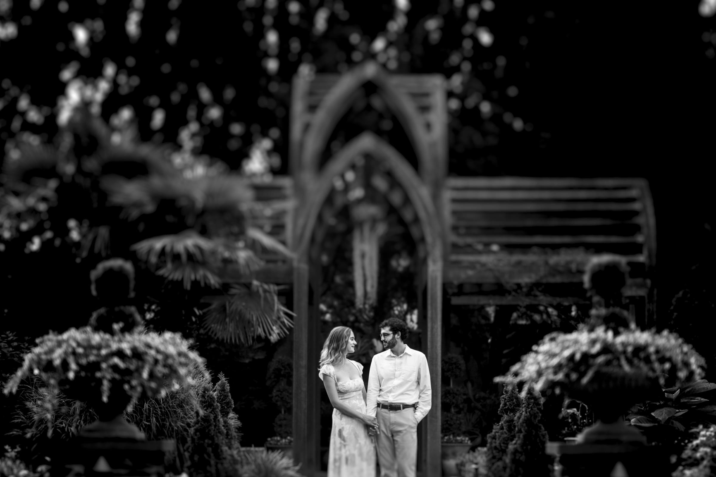 A black and white image captures a couple standing in Duke Gardens, smiling at each other. Framed by an arched wooden structure, with lush foliage and ornate planters, it's a perfect engagement session moment.