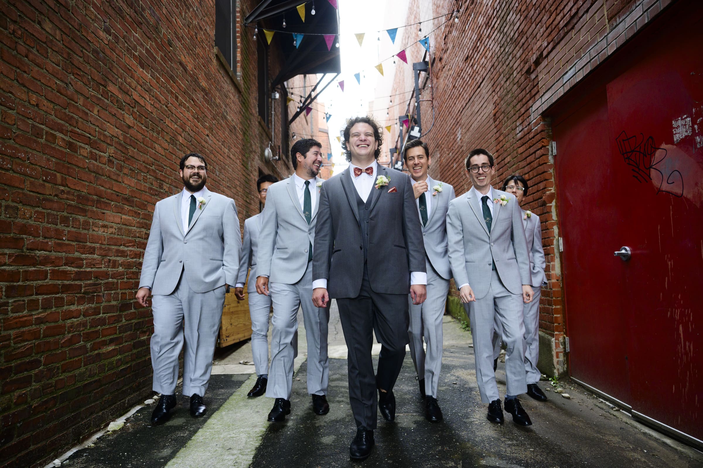 A group of groomsmen in gray suits and a groom in a dark suit stroll down an urban alleyway, fresh from their Duke Chapel Wedding. They are smiling and laughing under colorful pennant banners strung above. The brick alley walls add charm, with a striking red door on the right.
