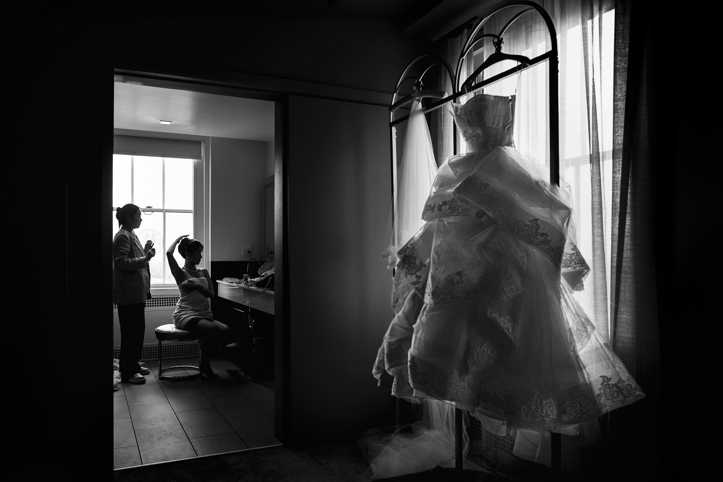 In a tranquil room before the Duke Chapel wedding, a bride sits gracefully as a stylist perfects her hair and makeup. By the window, her lace wedding dress is bathed in soft natural light, adding to the serene and anticipatory atmosphere.