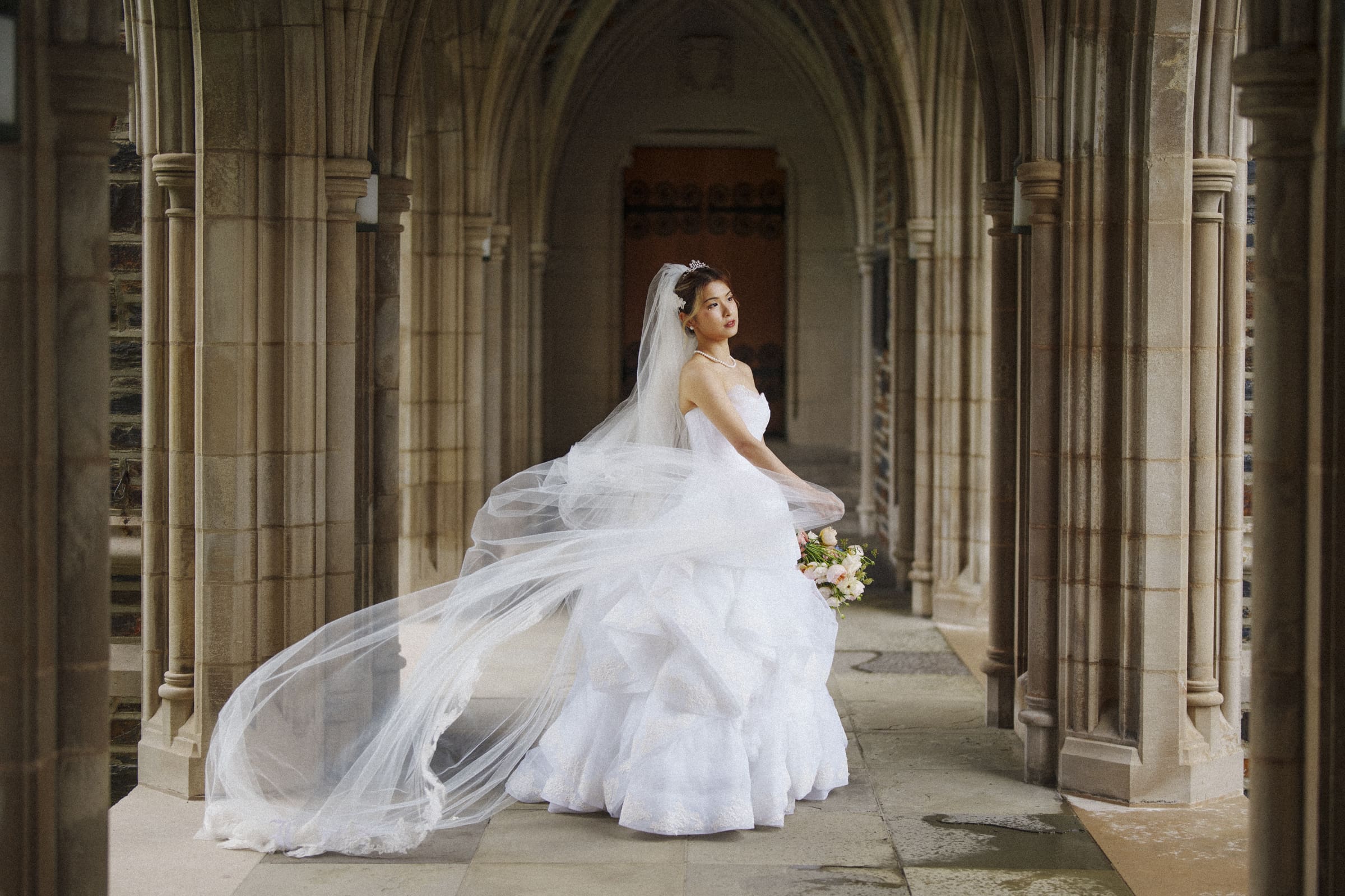 Duke Chapel Weddings An elegant bridal portrait of a Chinese bride in a white gown at Duke Chapel, looking away as the wind catches her long veil while she stands in a stone archway holding her bouquet.
