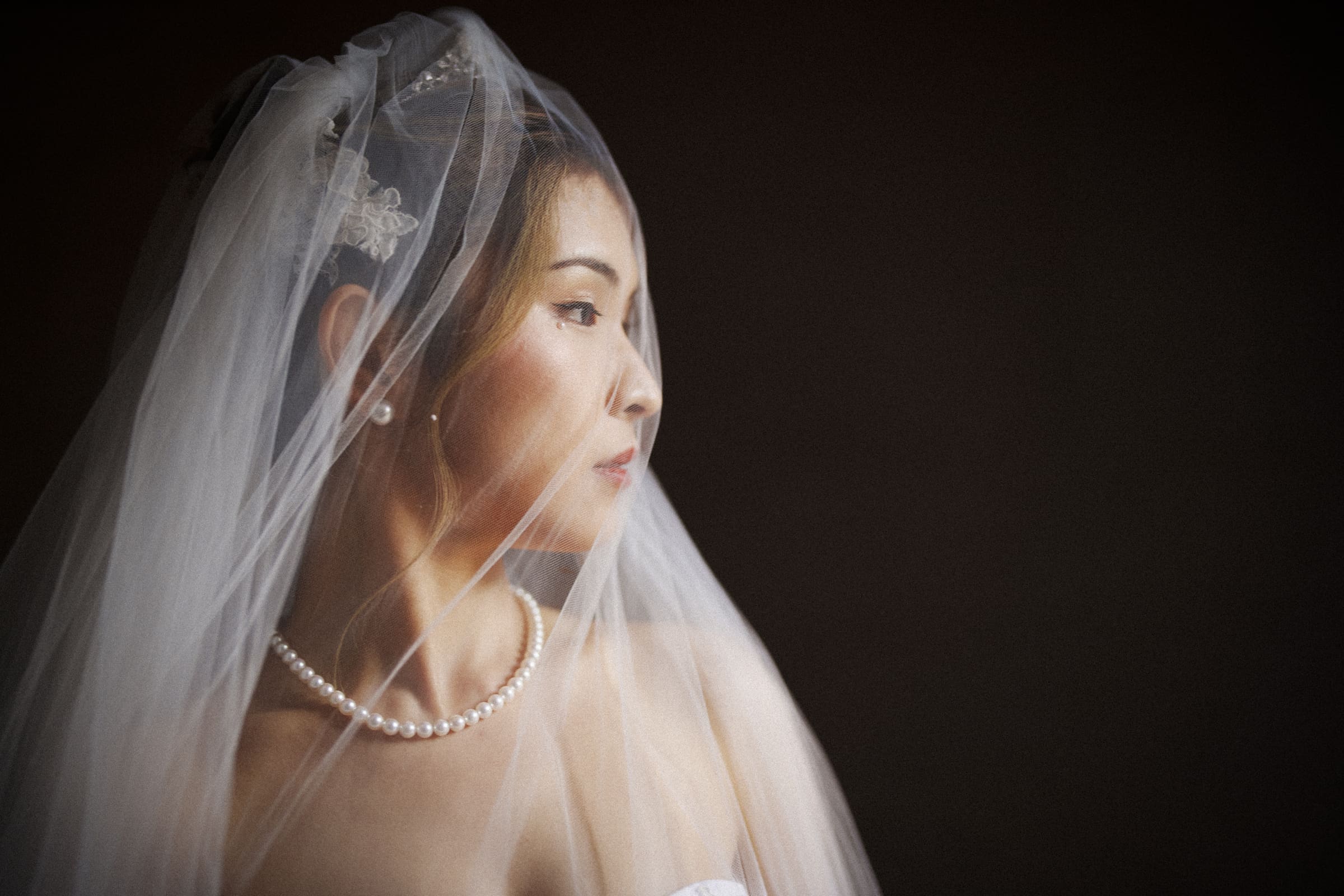 Duke Chapel Weddings A moody colour bridal portrait of a Chinese bride wearing a pearl necklace and white veil, looking to the side in a serene, contemplative pose against a dark background at Duke Chapel.