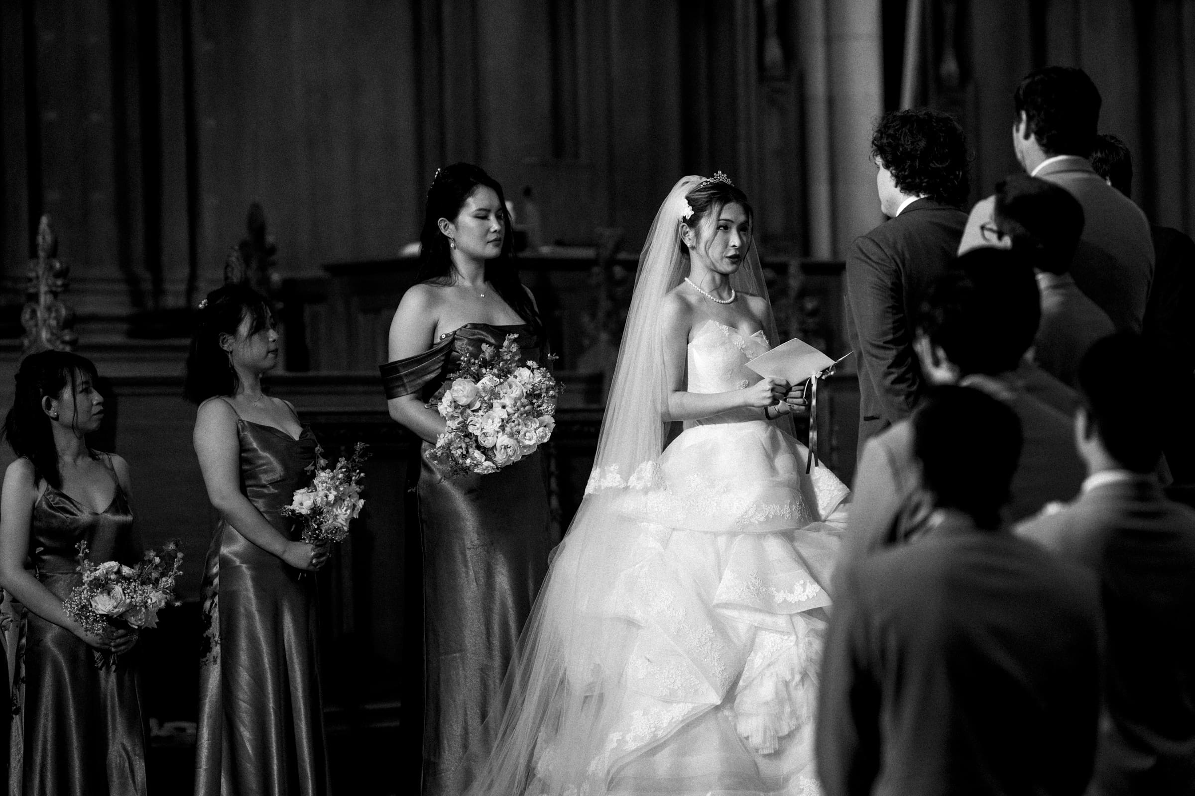 An emotional black and white wedding photograph inside Duke Chapel, capturing the bride reading her vows from a paper at the altar while the groom, bridesmaids, and guests look on.