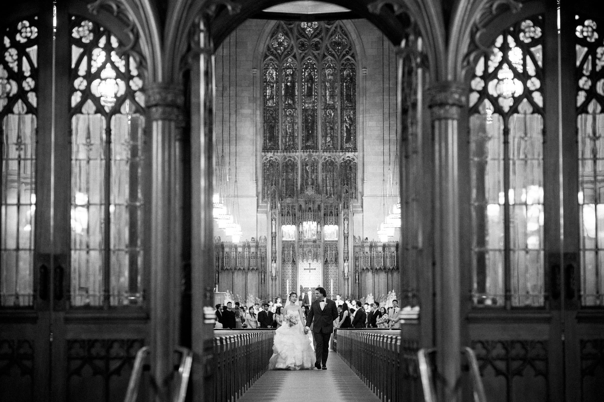 A bride and groom walk down the aisle of a grand, ornate church filled with seated guests, viewed through decorative archways in black and white during a beautiful Duke Chapel Wedding.