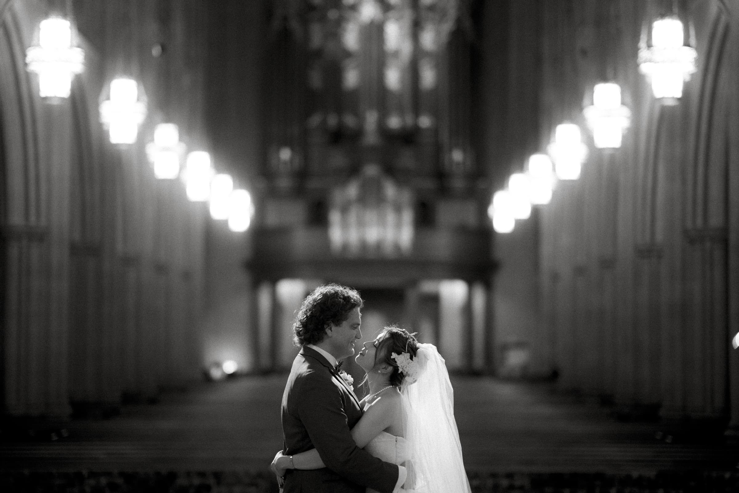 A couple in wedding attire embrace and smile at each other in the aisle of the grand Duke Chapel. Rows of glowing chandeliers illuminate the ornate, arched interior in this dimly lit, black and white photograph.