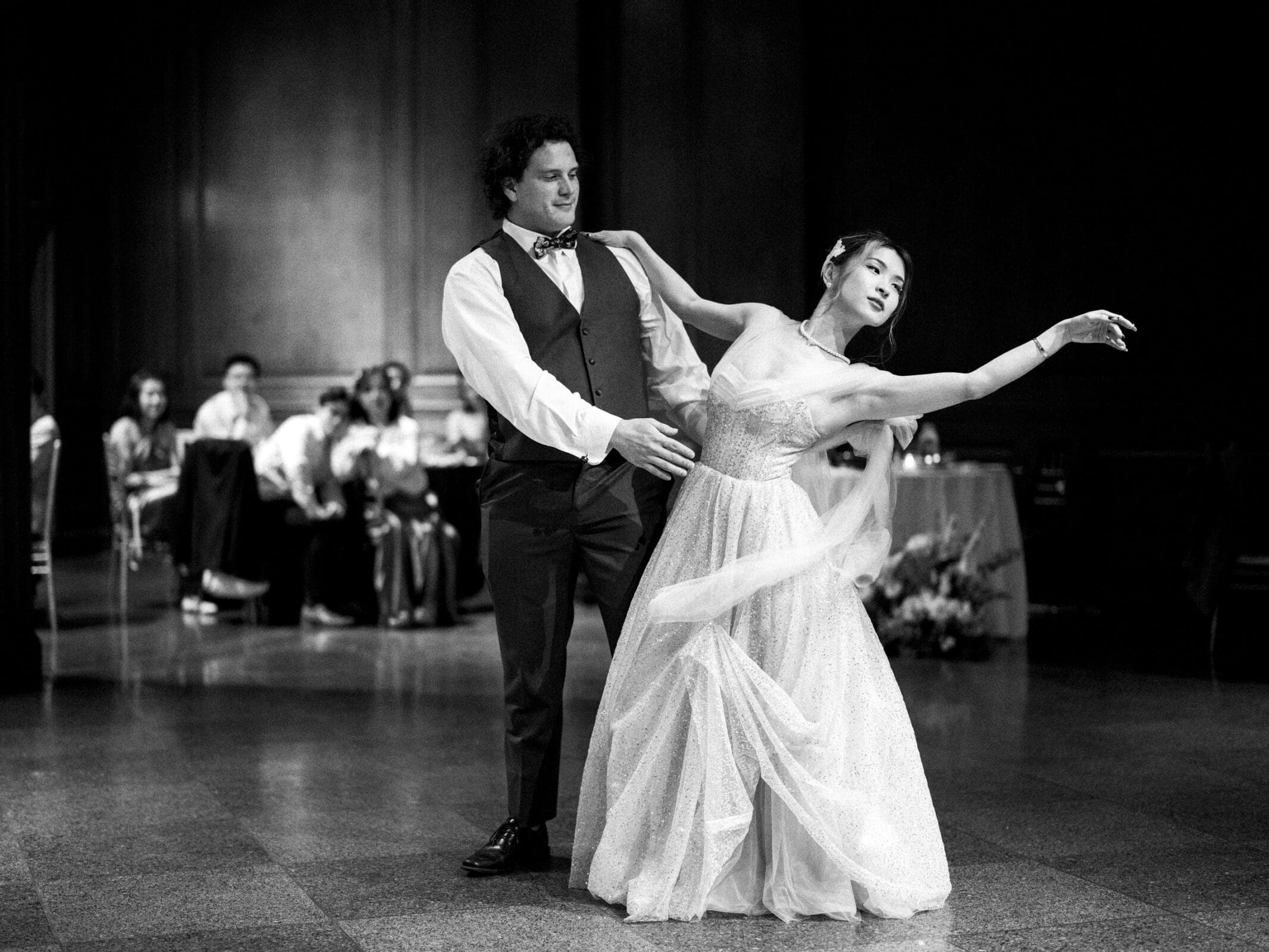 A couple elegantly dancing in formal attire at a Duke Chapel wedding, set in a dimly lit room with onlookers seated in the background. The woman, in a flowing dress, is gracefully extended while the man supports her, both appearing poised and composed.