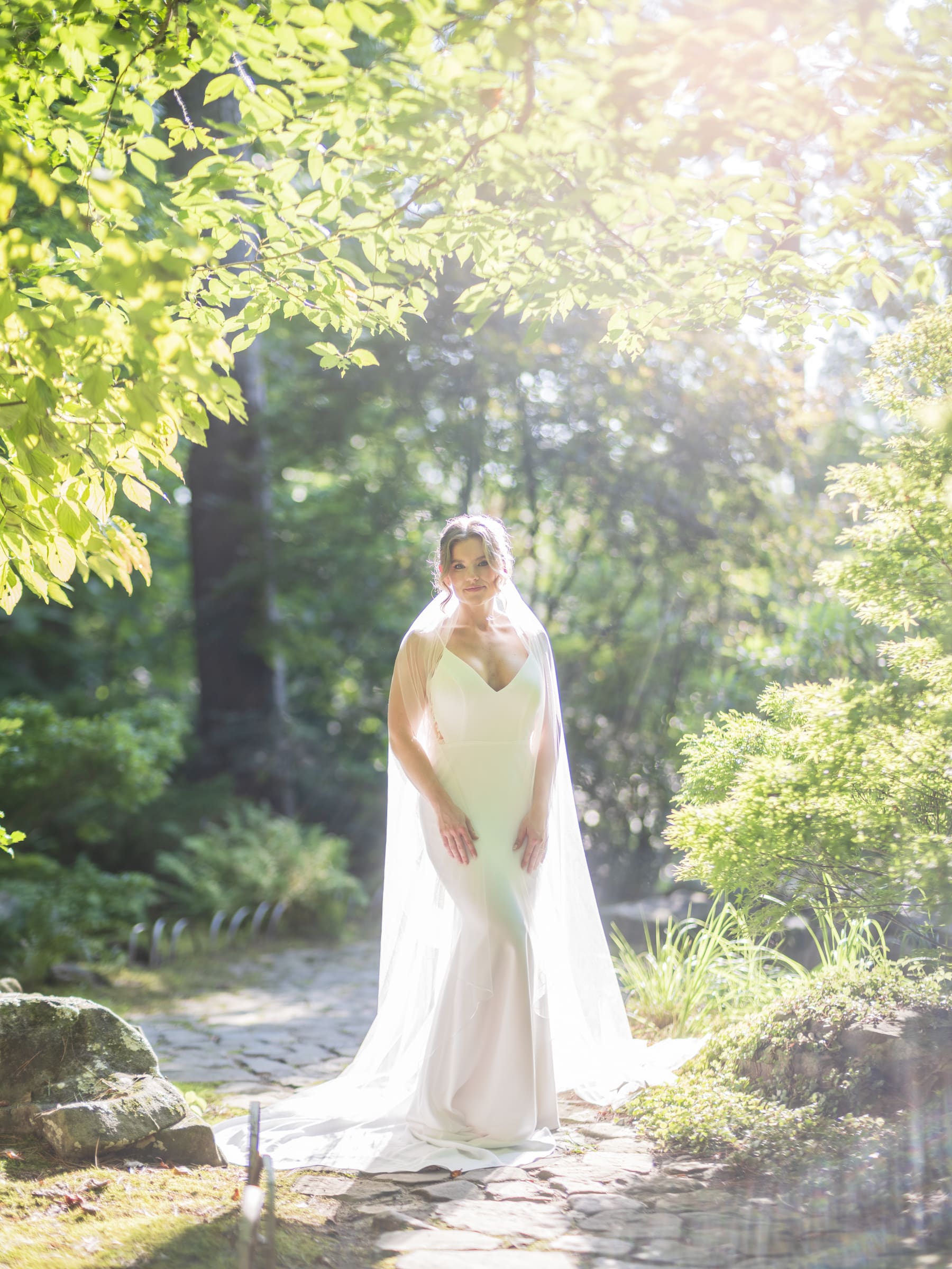 A woman in a white wedding dress and veil stands on a sunlit stone path surrounded by lush green trees and plants, sunlight filtering through the leaves—perfect for Duke Gardens photography.