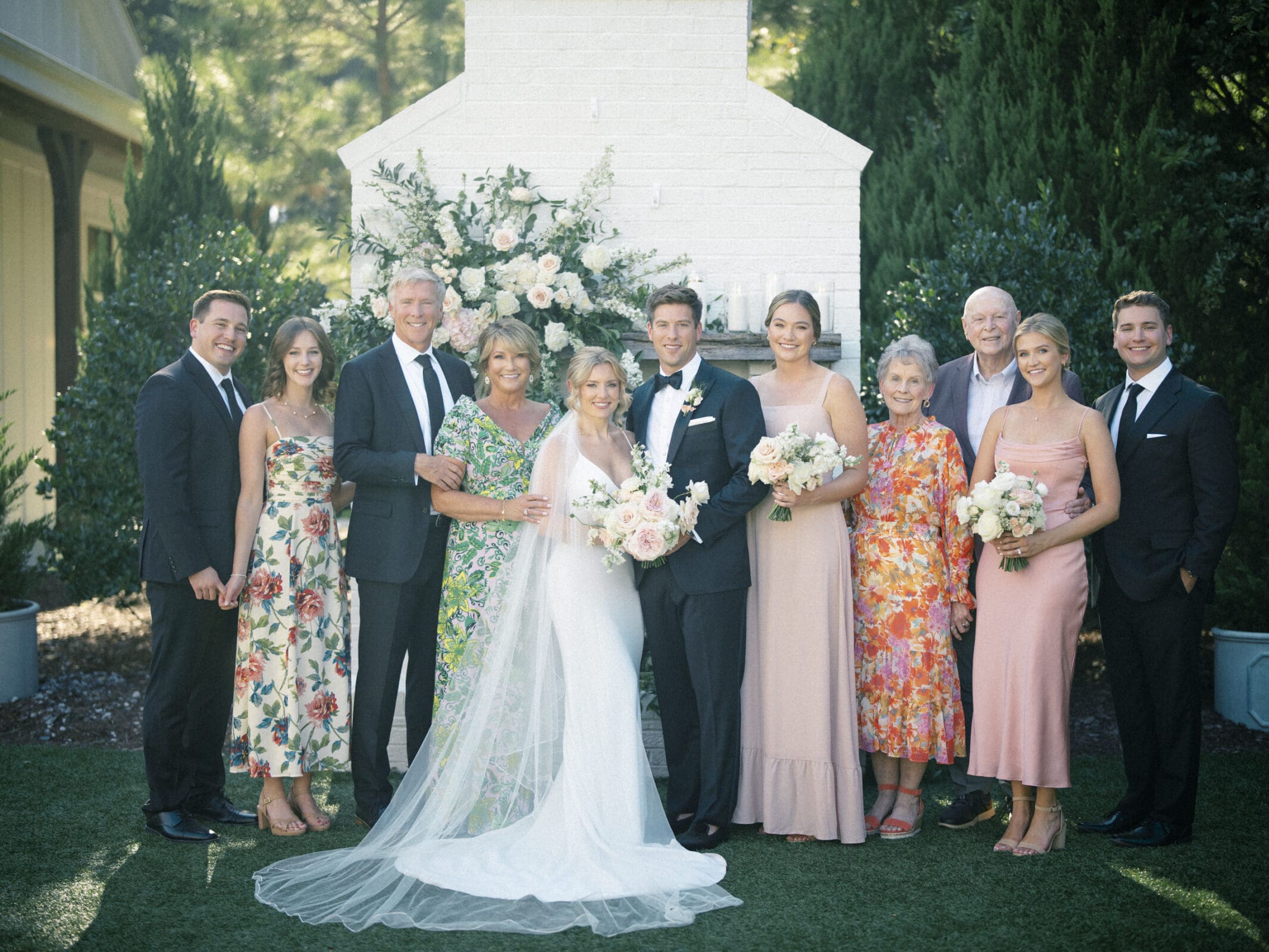 The Bradford Wedding group photo captures a bride and groom at the center, surrounded by family and friends. The bride, in a white gown with a bouquet, and the groom, in a black tuxedo, are framed by loved ones in vibrant attire against a backdrop of lush greenery.