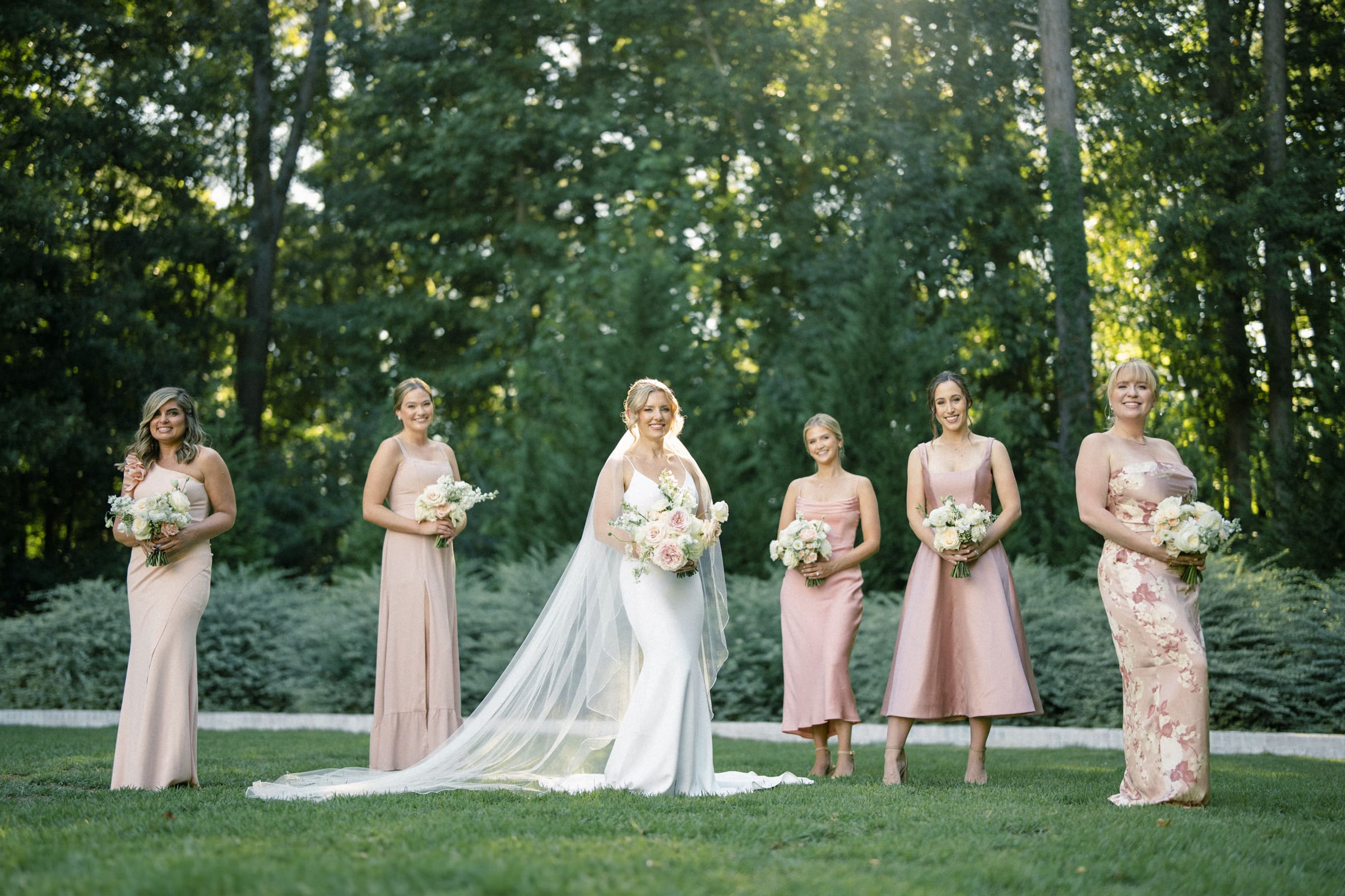 At The Bradford Wedding, a bride in a white dress stands with five bridesmaids in pink dresses, each holding bouquets of flowers. They are outside on a grassy area with trees in the background, under a soft, sunlit sky.
