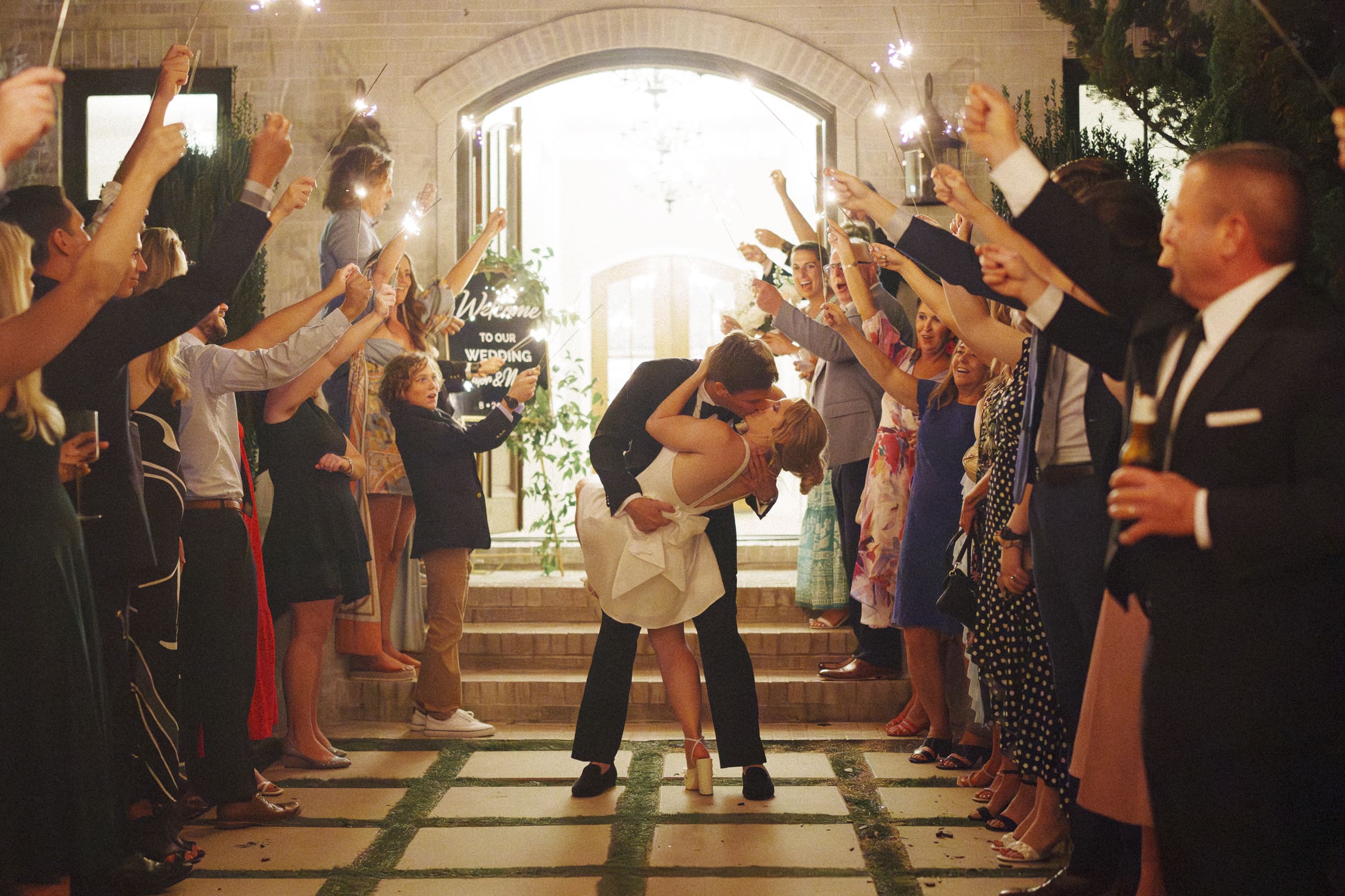 A couple kisses passionately under sparklers during their Bradford Wedding exit. Guests stand on either side, celebrating and cheering them on. The bride's white dress and the groom's suit contrast against the warmly lit scene.
