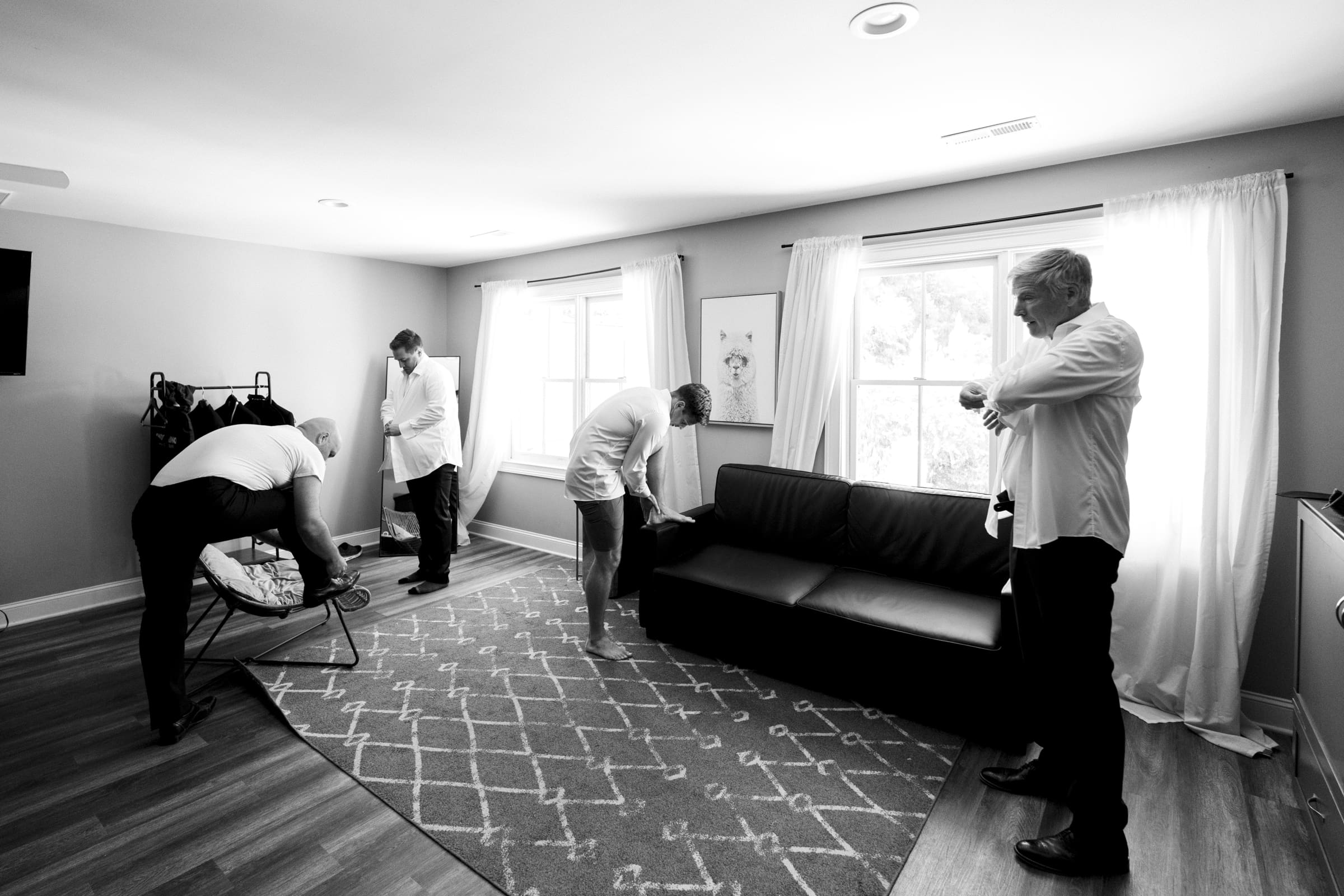 A black and white photo captures four men in a room at The Bradford Wedding, adjusting their clothing with meticulous care. Two are by the window, one bends over a chair, and another stands by the couch. The room features a patterned rug and light curtains, setting an elegant scene.