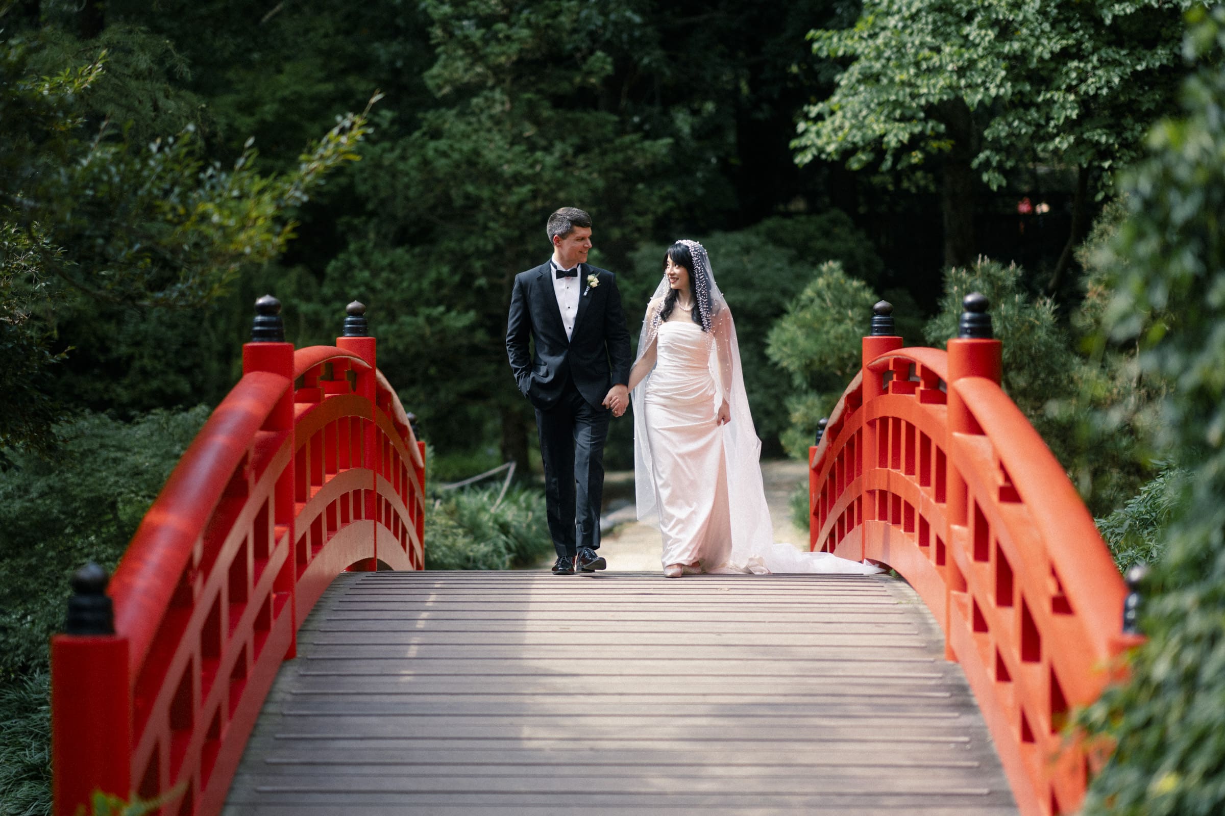 A bride and groom, fresh from their Duke Chapel wedding, walk hand in hand across a red arched wooden bridge in a lush green garden. She wears a white gown and veil, while he is in a black suit with a bow tie.