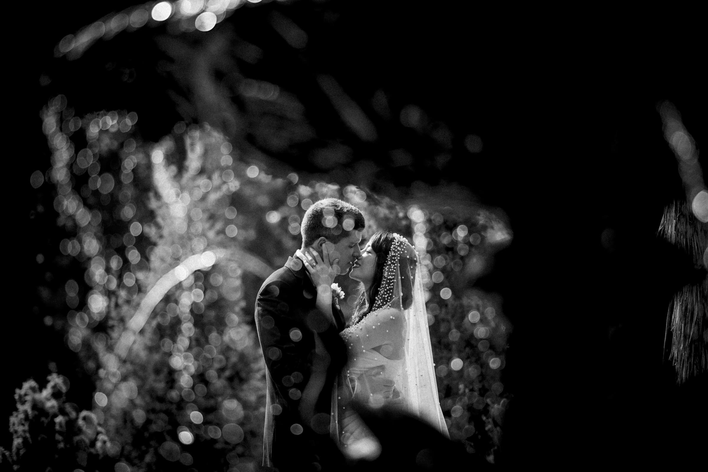 A black and white photo captures a bride and groom embracing amidst blurred bokeh lights and lush foliage at their Duke Chapel wedding. The bride, adorned with a delicate veil, leans into her groom, both dressed in formal attire as they share an intimate moment.