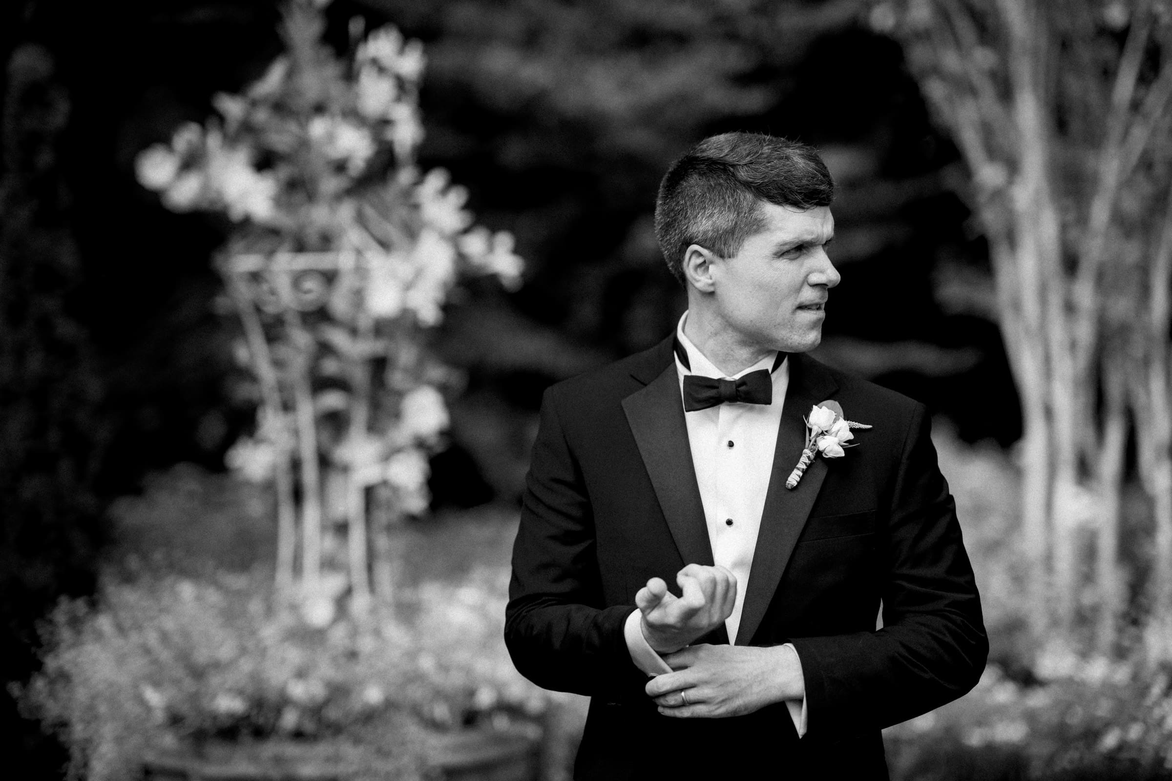 A man in a tuxedo with a boutonniere adjusts his cufflinks, evoking the elegance of a Duke Chapel wedding. The background features blurred foliage and floral arrangements. The black-and-white image enhances the classic and formal atmosphere.