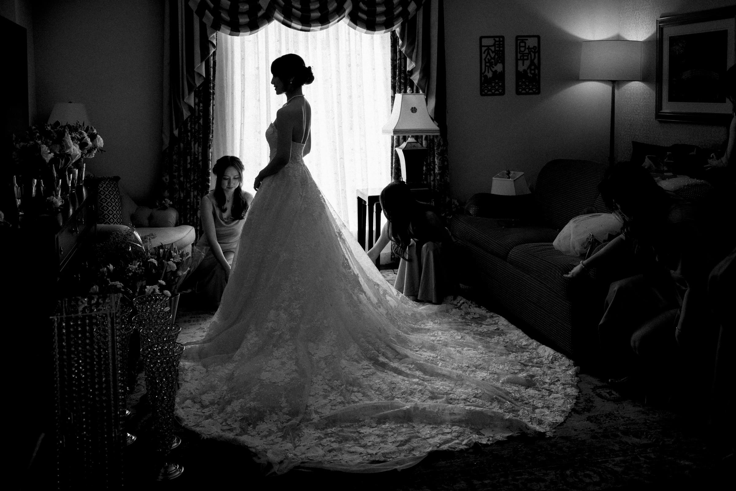A black and white photo captures the essence of a Duke Chapel wedding, as a bride in an elegant lace gown stands by a bright window. Her bridesmaids kneel nearby, adjusting her dress. The softly lit room, with patterned curtains and a cozy sofa in the background, exudes timeless charm.