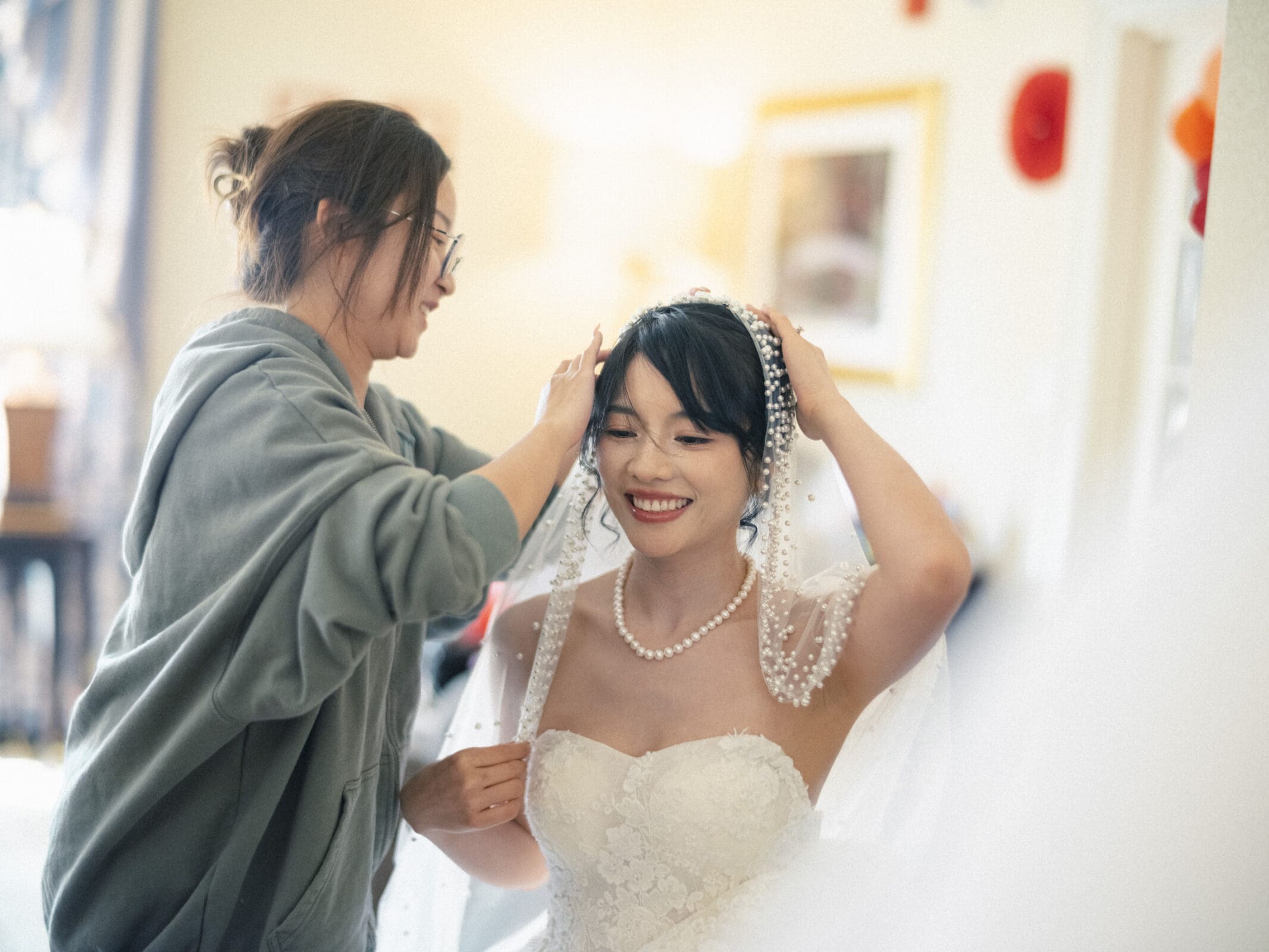 A bride in a strapless wedding dress and pearl necklace smiles as another woman adjusts her veil. The setting is indoors, with warm lighting and soft-focus elements in the background.