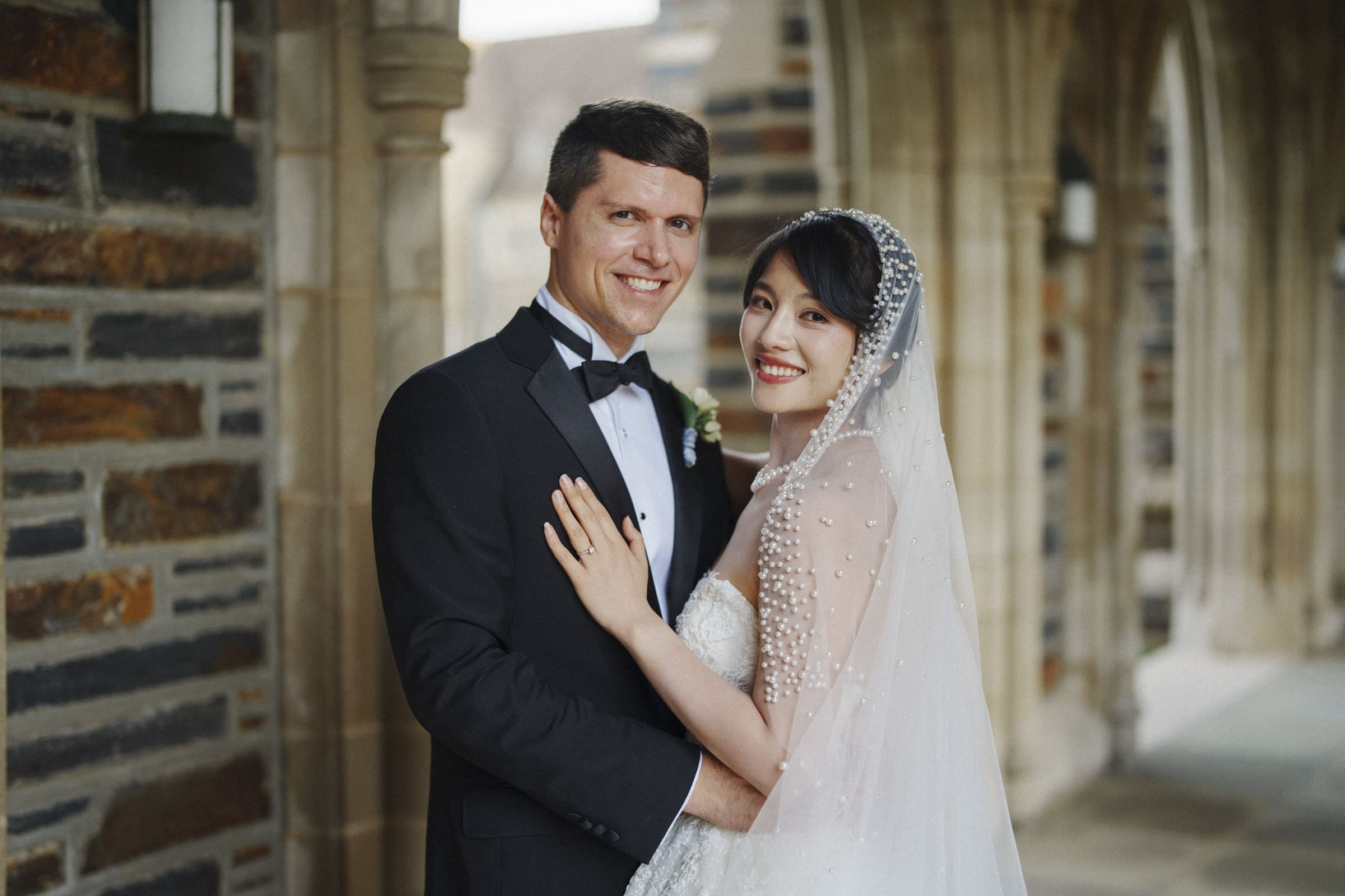 Duke Chapel Weddings A classic wedding portrait of a bride and groom smiling at the camera in the stone arcades of Duke Chapel, showcasing her white gown and beaded veil alongside his black tuxedo.