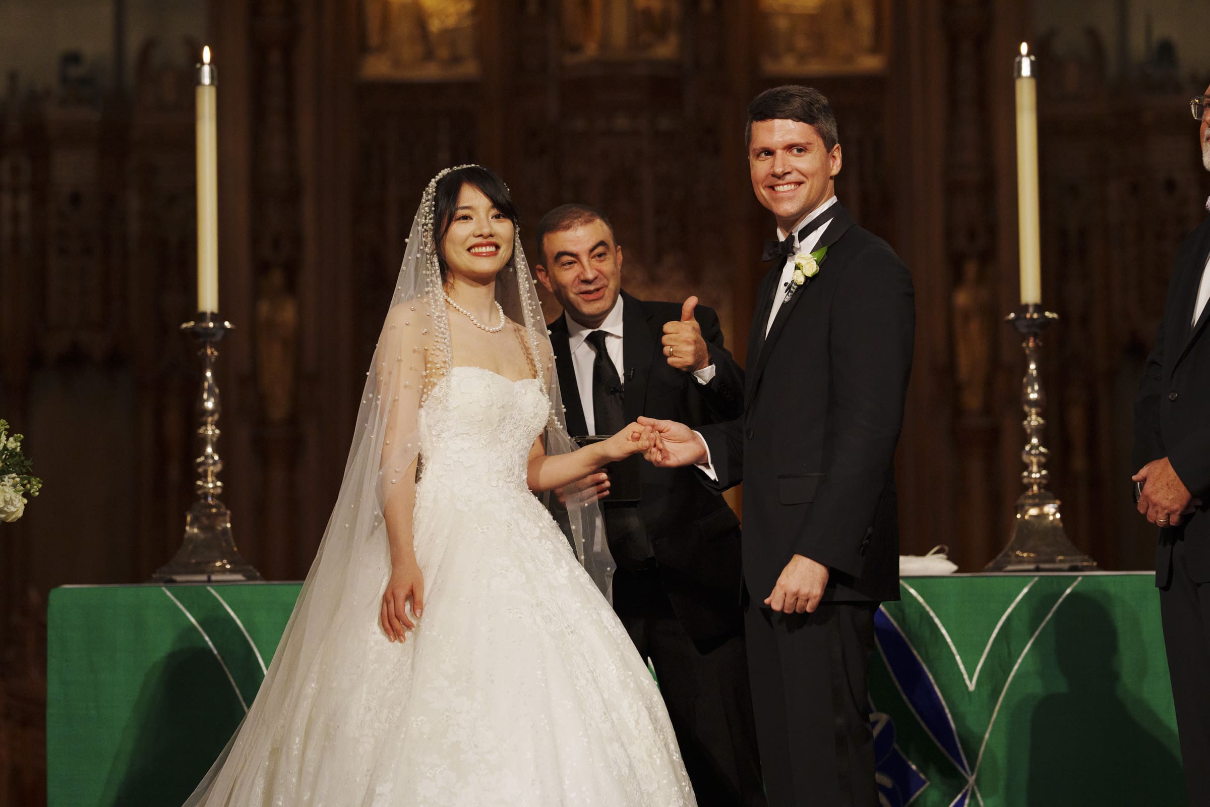 Duke Chapel Weddings A joyful, candid wedding photograph at the Duke Chapel altar, showing the bride and groom smiling and holding hands while the officiant next to them gives a thumbs-up by two tall candles