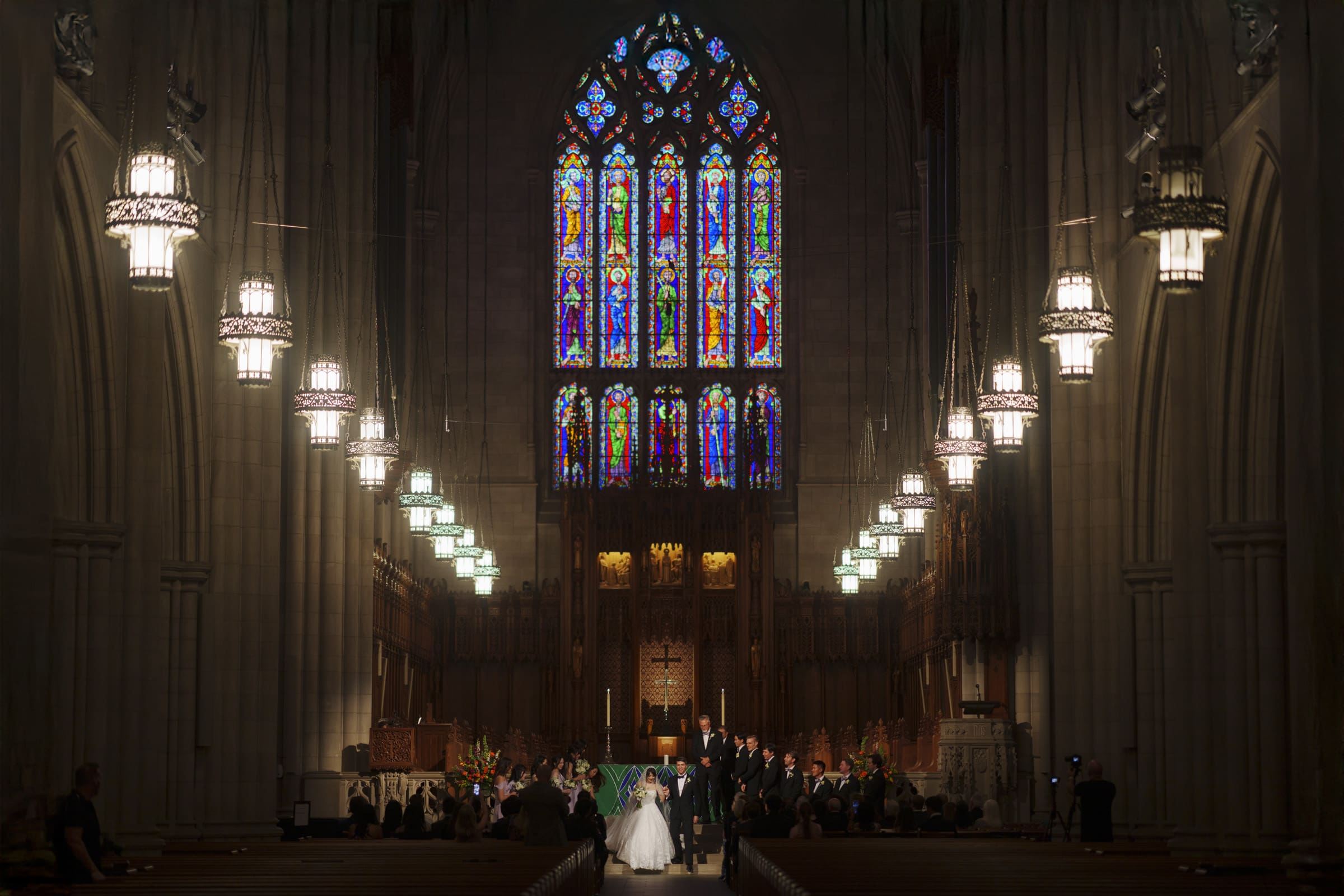 A couple stands at the altar in a grand cathedral with high ceilings. Stained glass windows above cast colorful light into the dimly lit space. Large chandeliers and wooden pews line the aisle, creating an elegant atmosphere for the wedding ceremony.