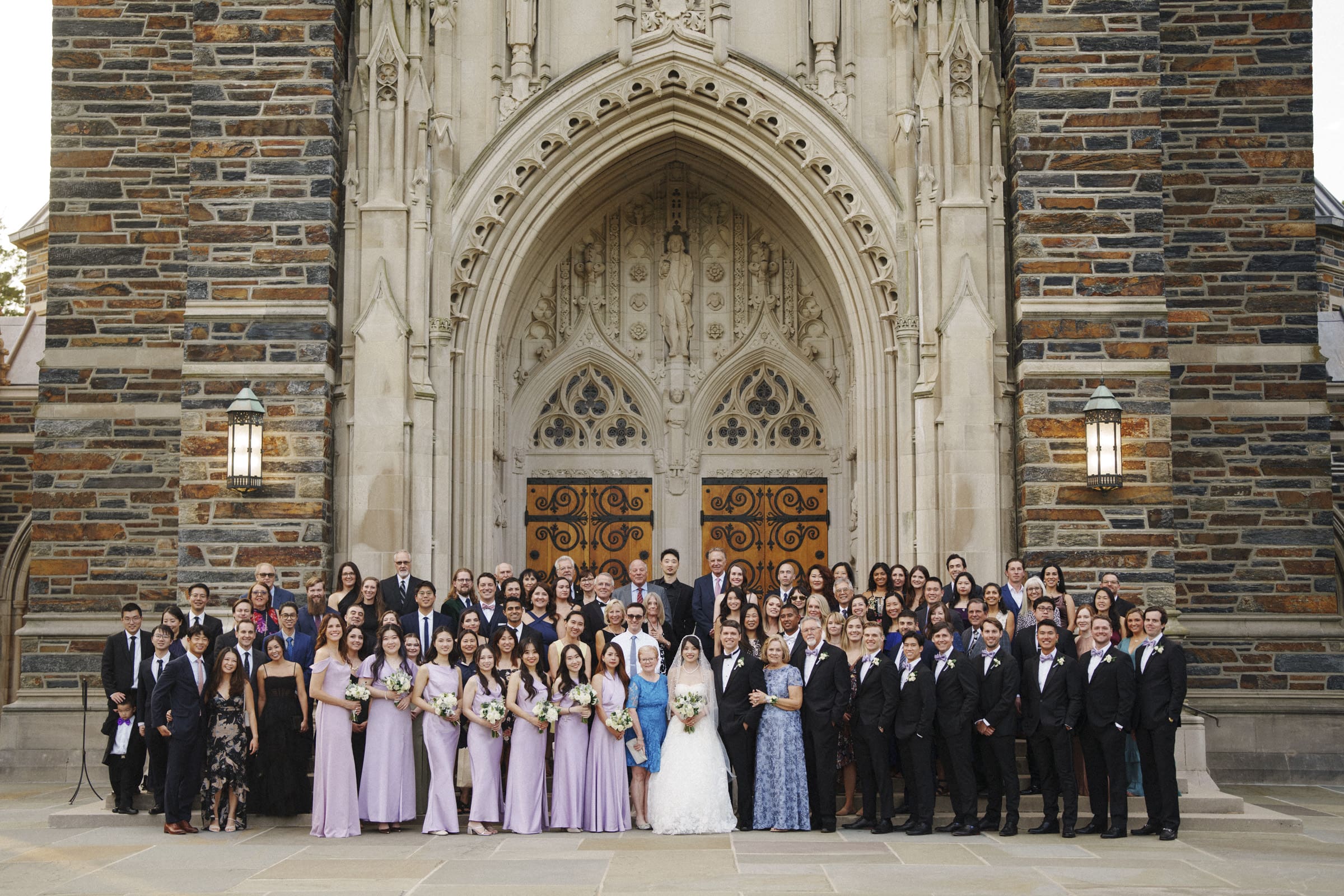 A large wedding group poses in front of Duke Chapel. entrance. The bride and groom stand at the center, surrounded by bridesmaids in lavender dresses and groomsmen in suits, along with other guests. The church features ornate stonework and large wooden doors.