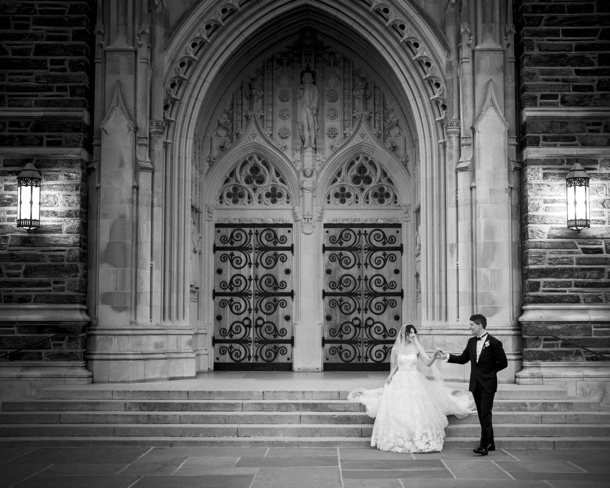 A classic black and white wedding photograph of a groom holding his bride's hand to help her walk down the grand stone steps of the Duke Chapel entrance, illuminated by glowing lanterns.