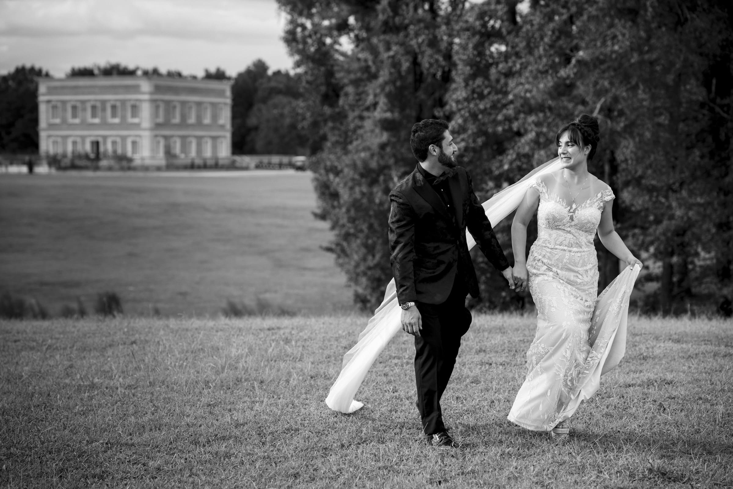 A bride and groom walk hand in hand across a field, smiling at each other, captured beautifully by Oxbow Estate Wedding Photography. The bride wears a flowing dress with a train, and the groom is in a dark suit. A large building is visible in the background, surrounded by trees.