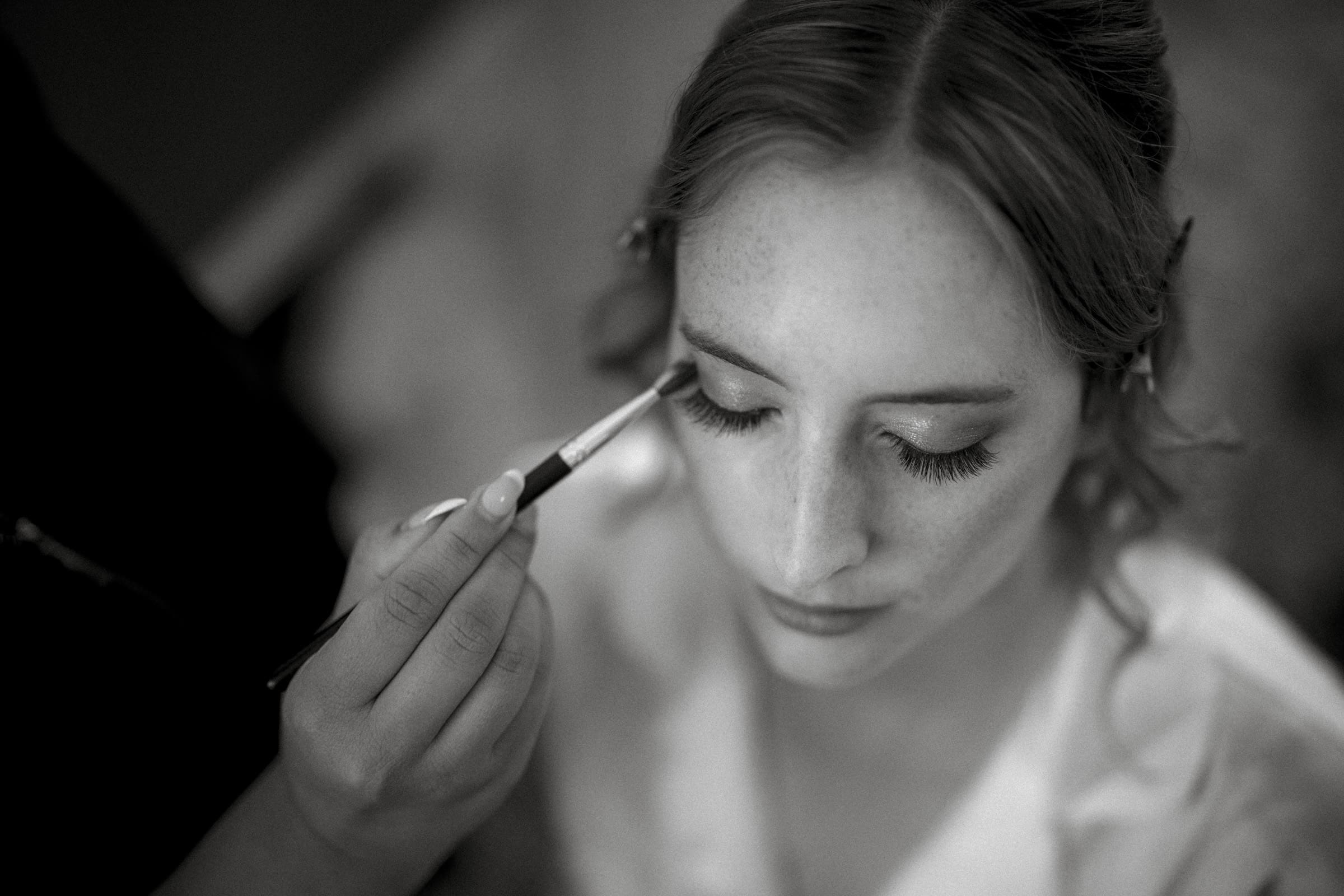 A black and white image of a person with closed eyes having makeup applied with a brush. Hair is pinned back, and the focus is on their serene expression and the makeup artists hand.