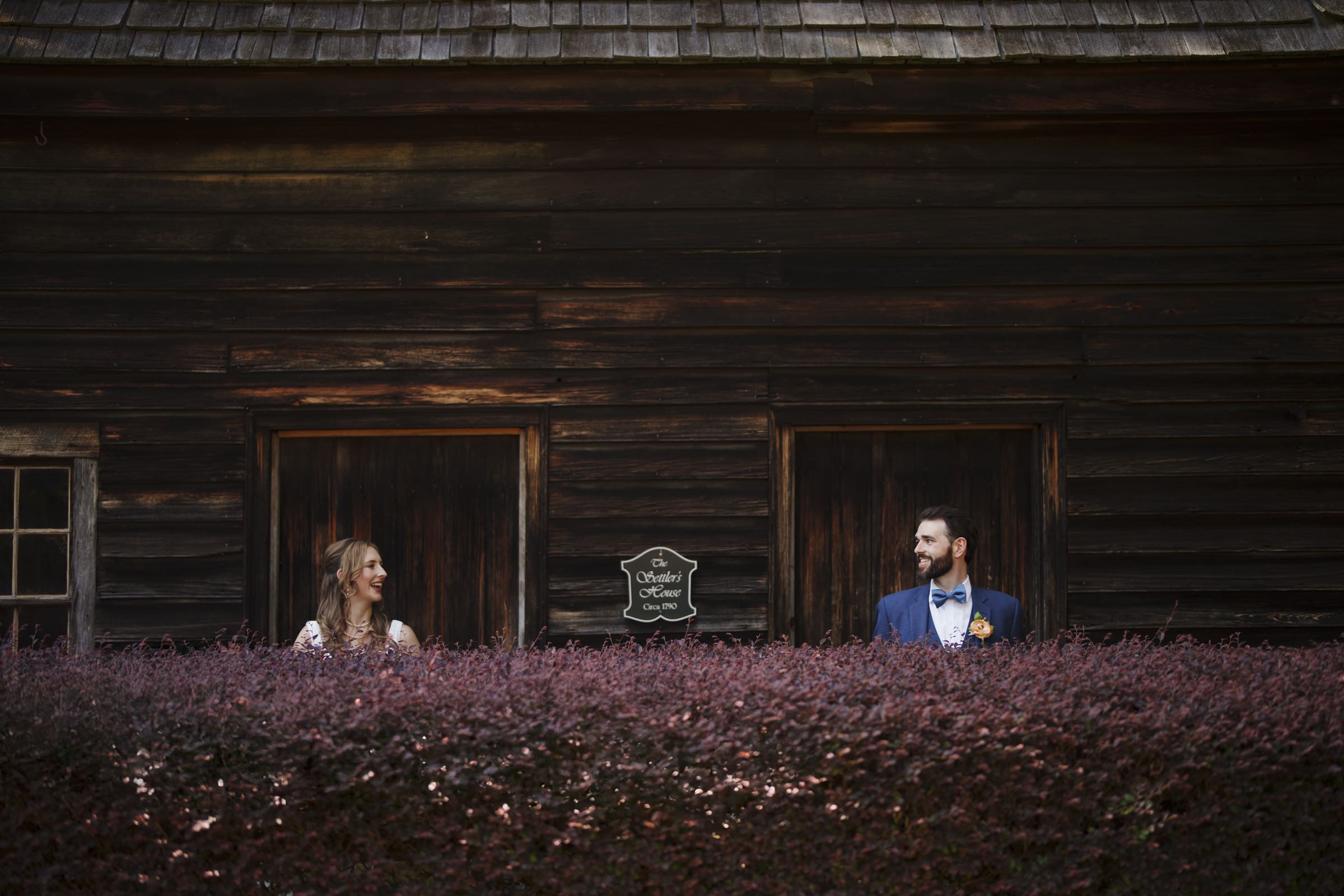 A bride in a white dress and a groom in a blue suit with a bow tie stand in separate wooden doorways of an old building. They are looking at each other over a bush with purple leaves. A sign between the doors reads School House.