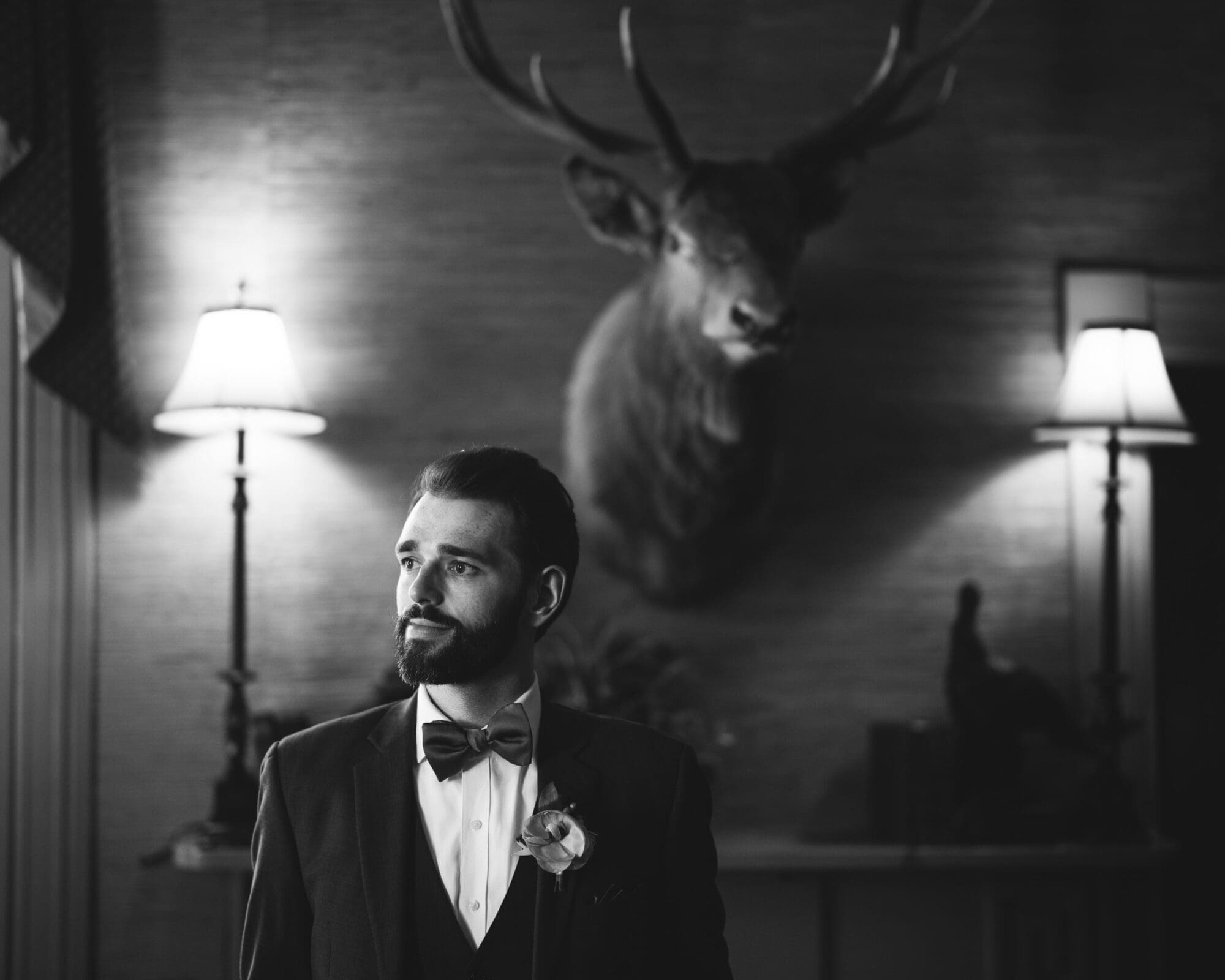 A groom with a beard stands in a room, wearing a suit with a bow tie and boutonniere. Two lamps flank him, and a mounted deer head is visible on the wall behind. The image is in black and white.