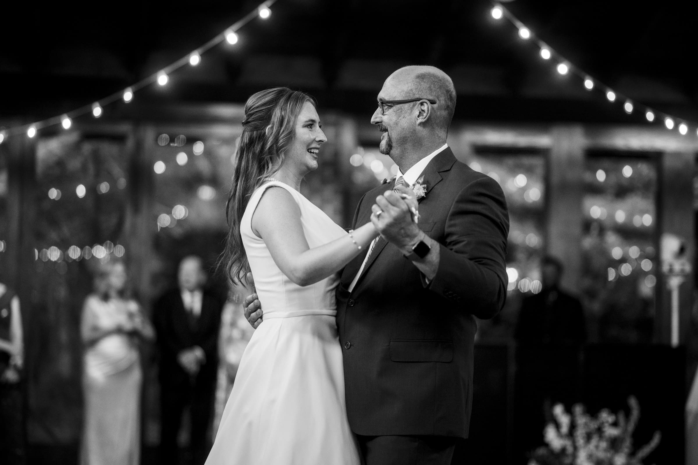 A black and white photo of a wedding dance. A bride in a flowing dress and a man in a suit smile at each other under string lights. They are holding hands while dancing, with blurred guests in the background.