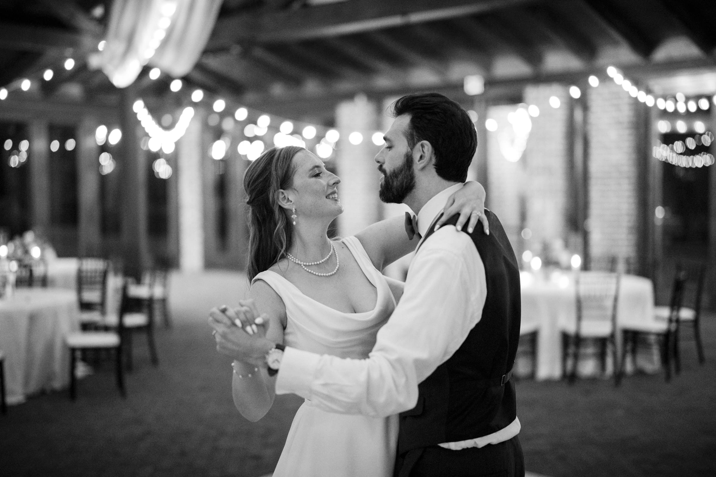 A couple elegantly dances together in a warmly lit room. The woman wears a white dress and pearl necklace, and the man is in a vest and tie. String lights create a romantic ambiance, with tables and chairs in the background.