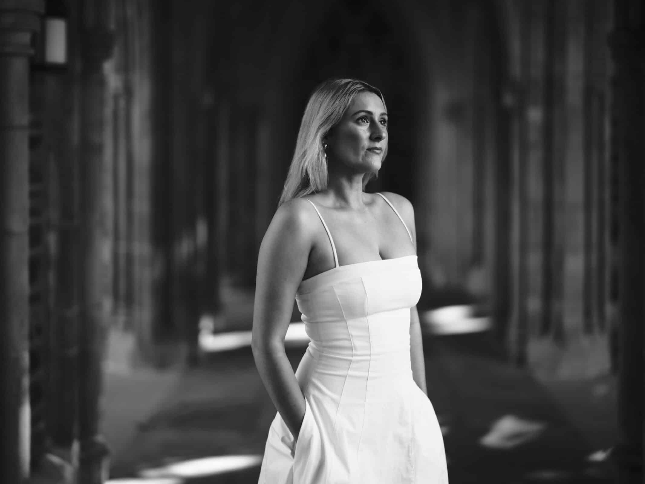 A woman in a sleeveless white dress stands in a stone corridor with her hands in her pockets, looking confidently into the distance. The photo is black and white, with soft natural light and arched columns in the background.