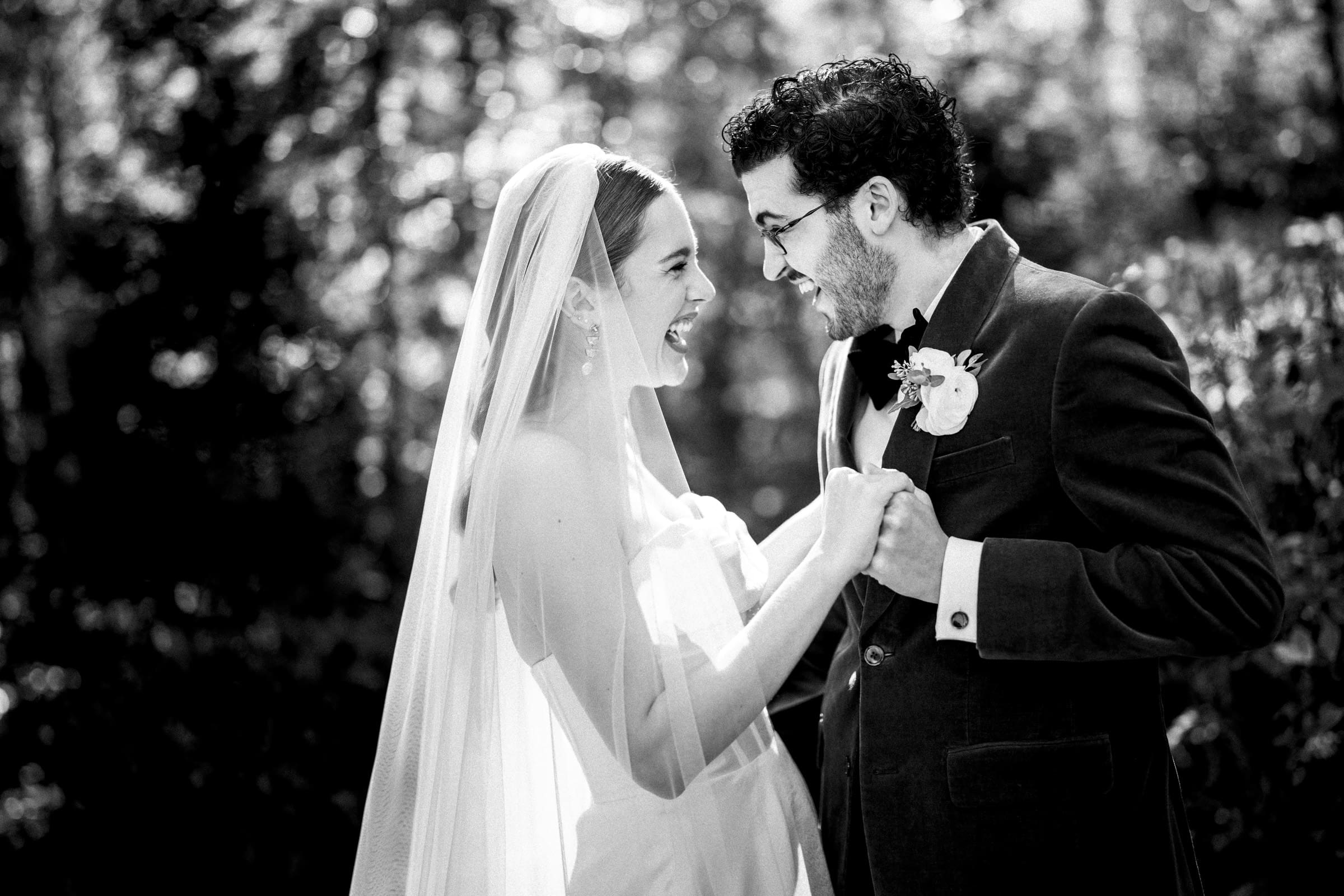 A bride and groom hold hands and smile joyfully at each other outdoors, surrounded by blurred trees and sunlight, both dressed in formal wedding attire.