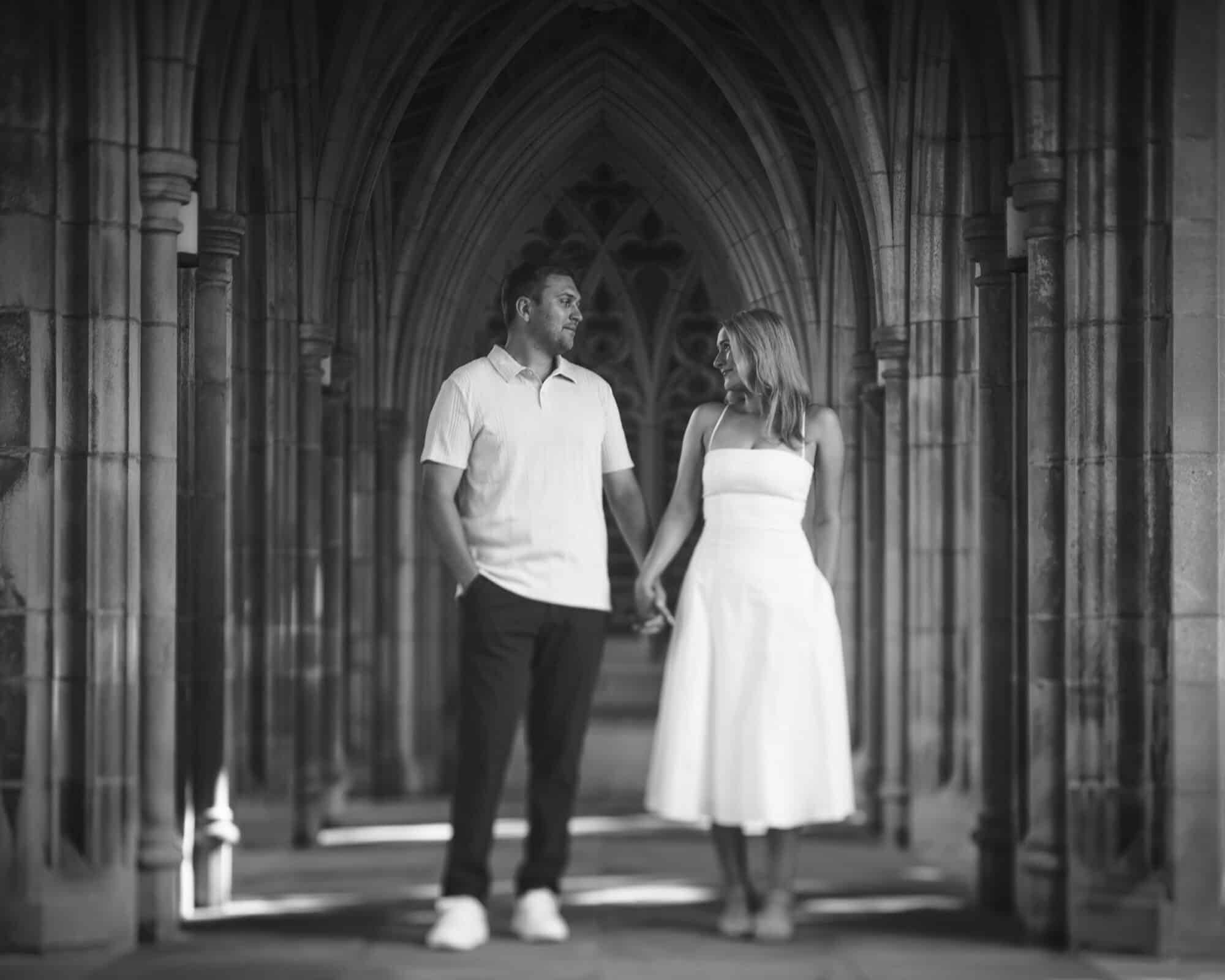 A couple holding hands and looking at each other stands in a stone corridor with arched ceilings. The image is in black and white, creating a romantic and timeless atmosphere.