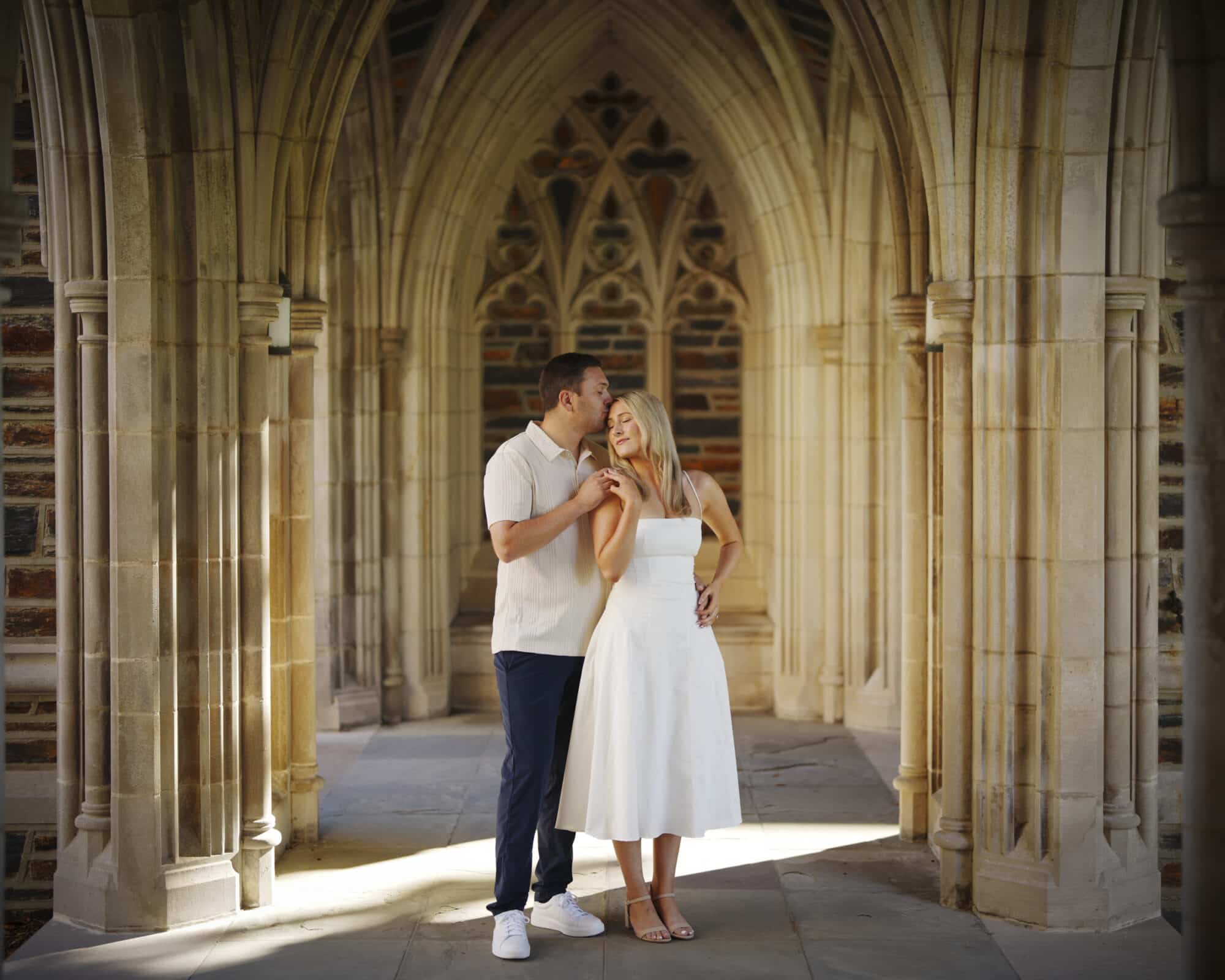 A couple stands closely under gothic stone arches, with the man kissing the woman’s forehead. The woman wears a white dress and sandals; the man wears a light shirt, dark pants, and sneakers. Sunlight filters through the arches.