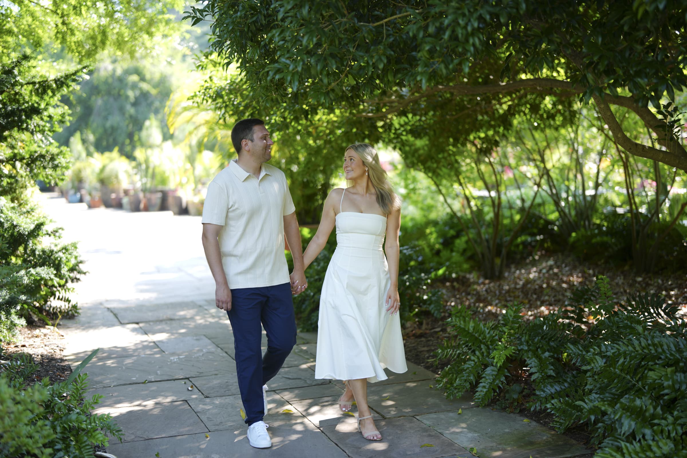 A man and woman walk hand in hand along a shaded garden path, surrounded by lush greenery. The man wears a light shirt and pants, while the woman wears a white dress. Both are smiling and looking at each other.