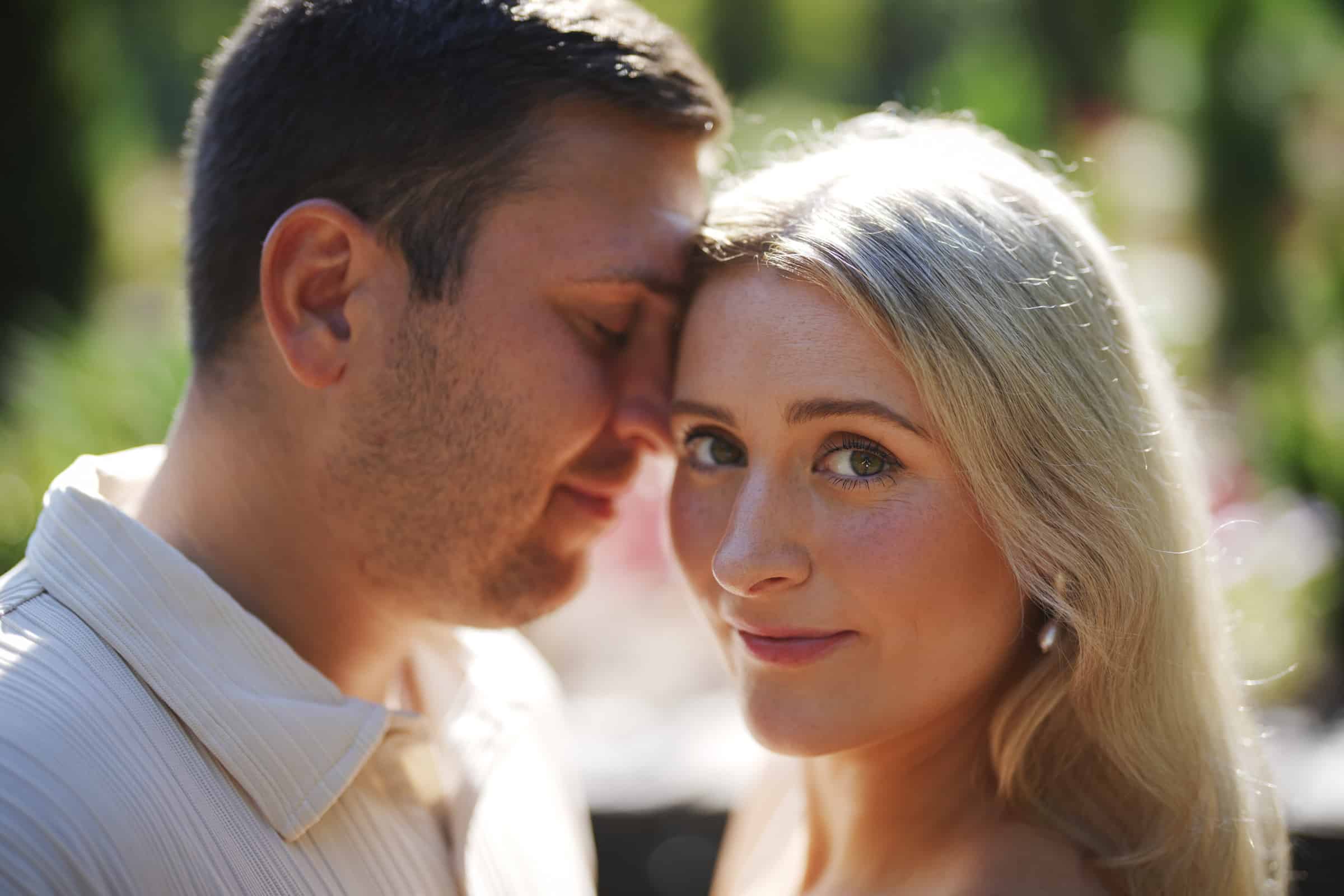 A man and a woman stand close together outdoors. The man has his eyes closed and leans his forehead toward the woman, while the woman with long blonde hair smiles softly at the camera. Sunlight filters through a blurred green background.