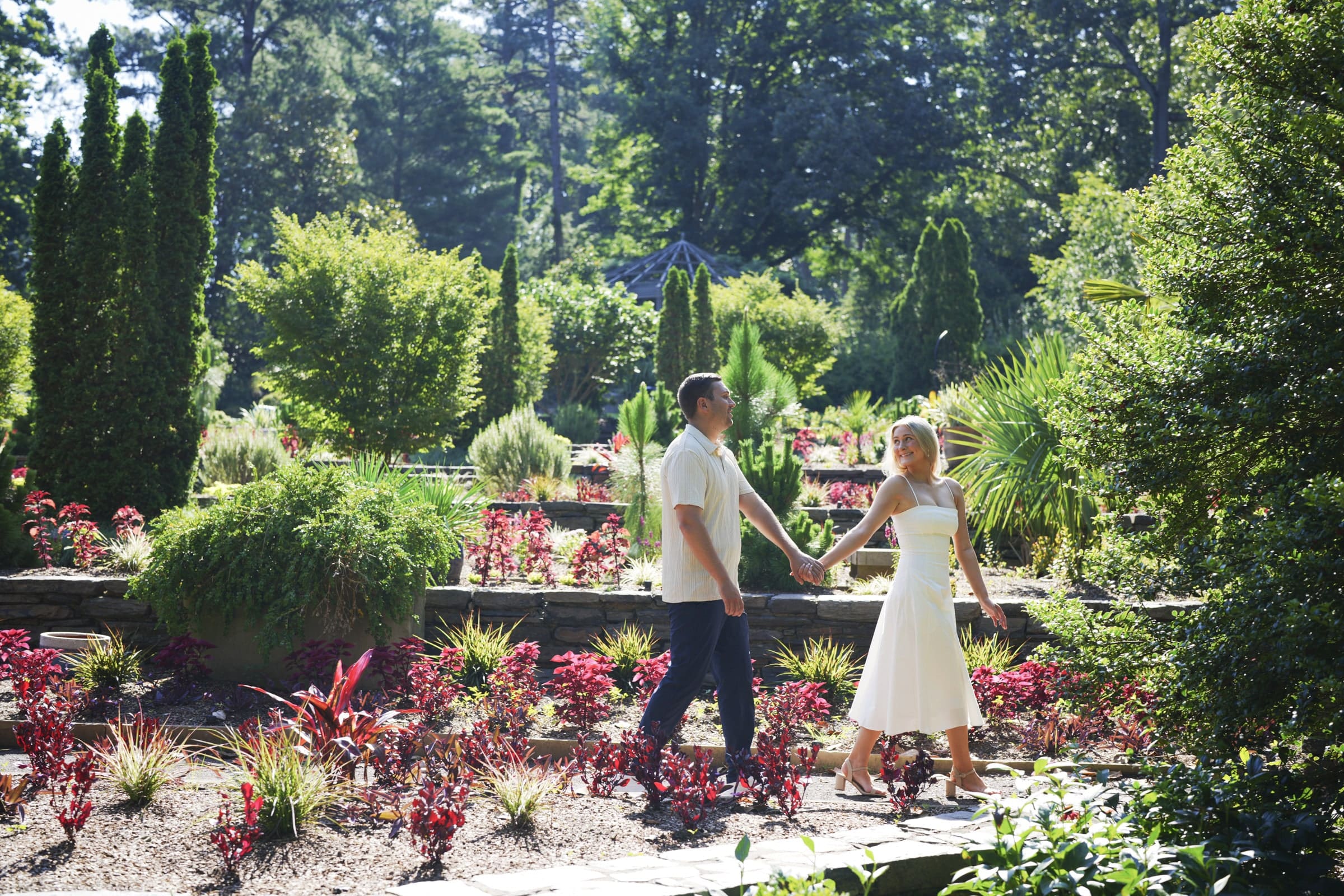 A couple holding hands and smiling as they walk along a path in a lush, sunlit botanical garden filled with vibrant green plants and colorful flowers.