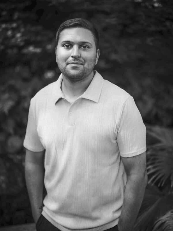 A man with short hair and a trimmed beard stands outdoors, wearing a light-colored, short-sleeved polo shirt. He has his hands in his pockets and gazes at the camera, with leafy plants blurred in the background.