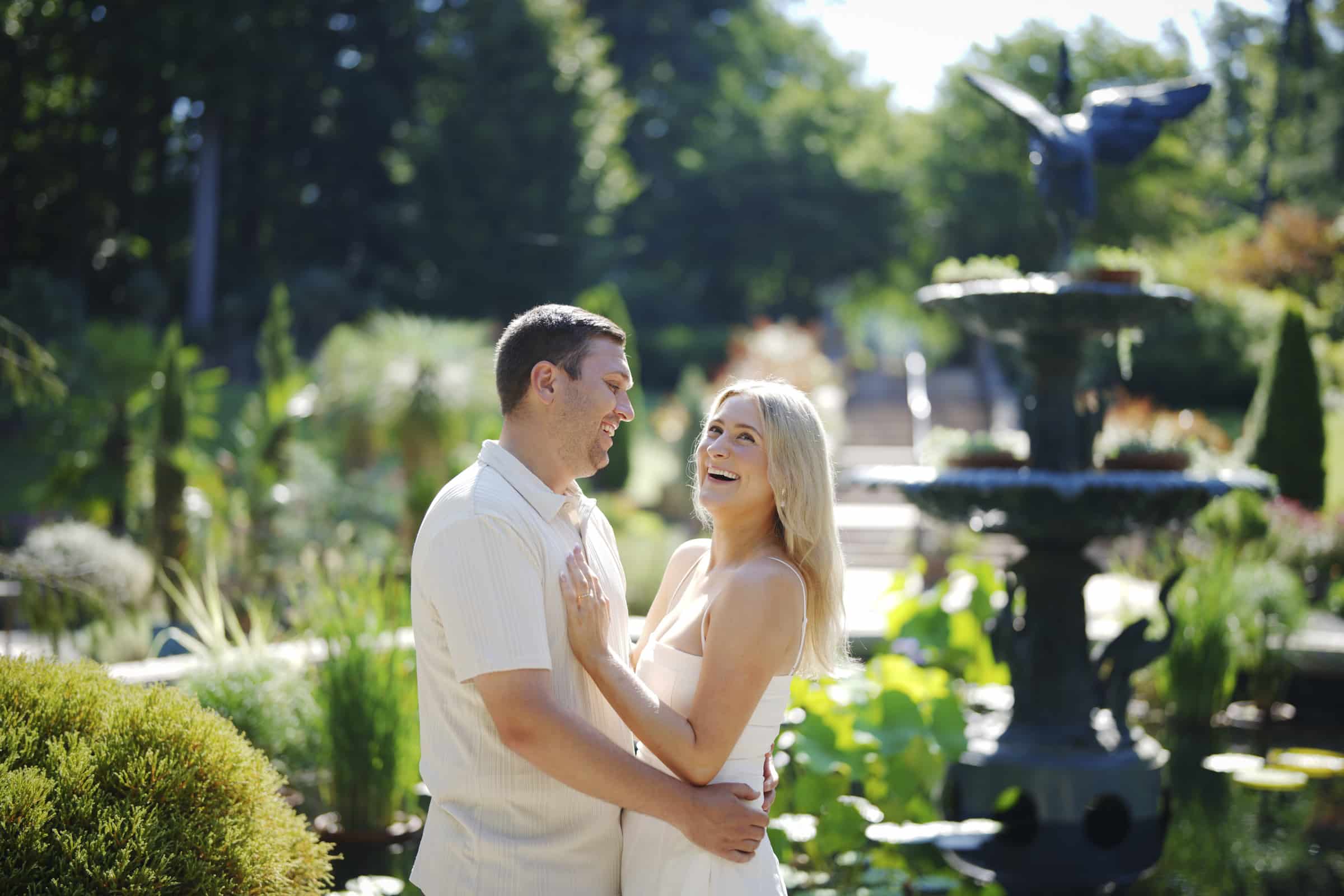A couple stands smiling and embracing in a sunlit garden near a large fountain, surrounded by greenery and blooming plants, on a bright, clear day.