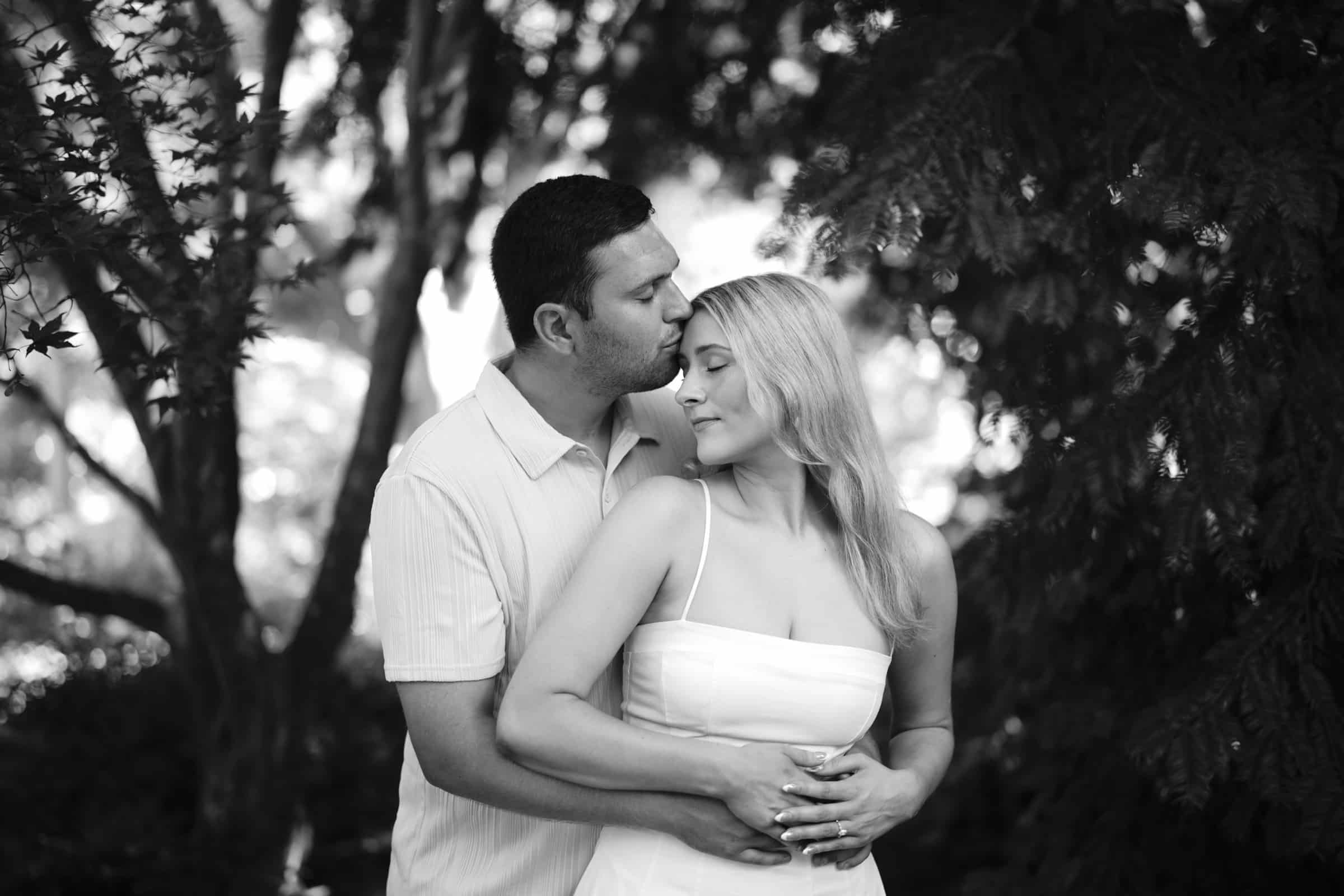 A man gently hugs a woman from behind and kisses her on the forehead as she smiles softly, both standing in a lush, sunlit garden. The photo is in black and white.