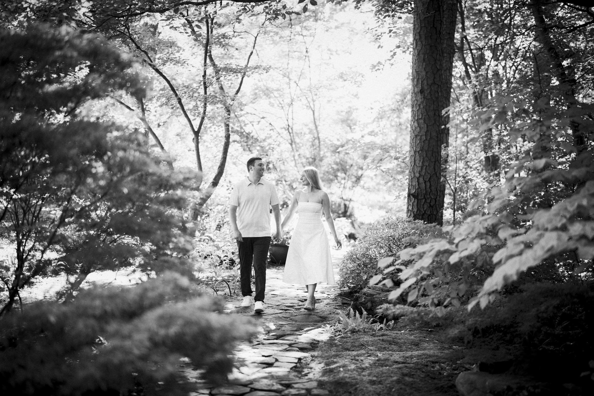 A couple walks hand-in-hand along a stone path in a lush, sunlit forest. Both are casually dressed and smiling, surrounded by trees and greenery in a peaceful, natural setting. The image is in black and white.