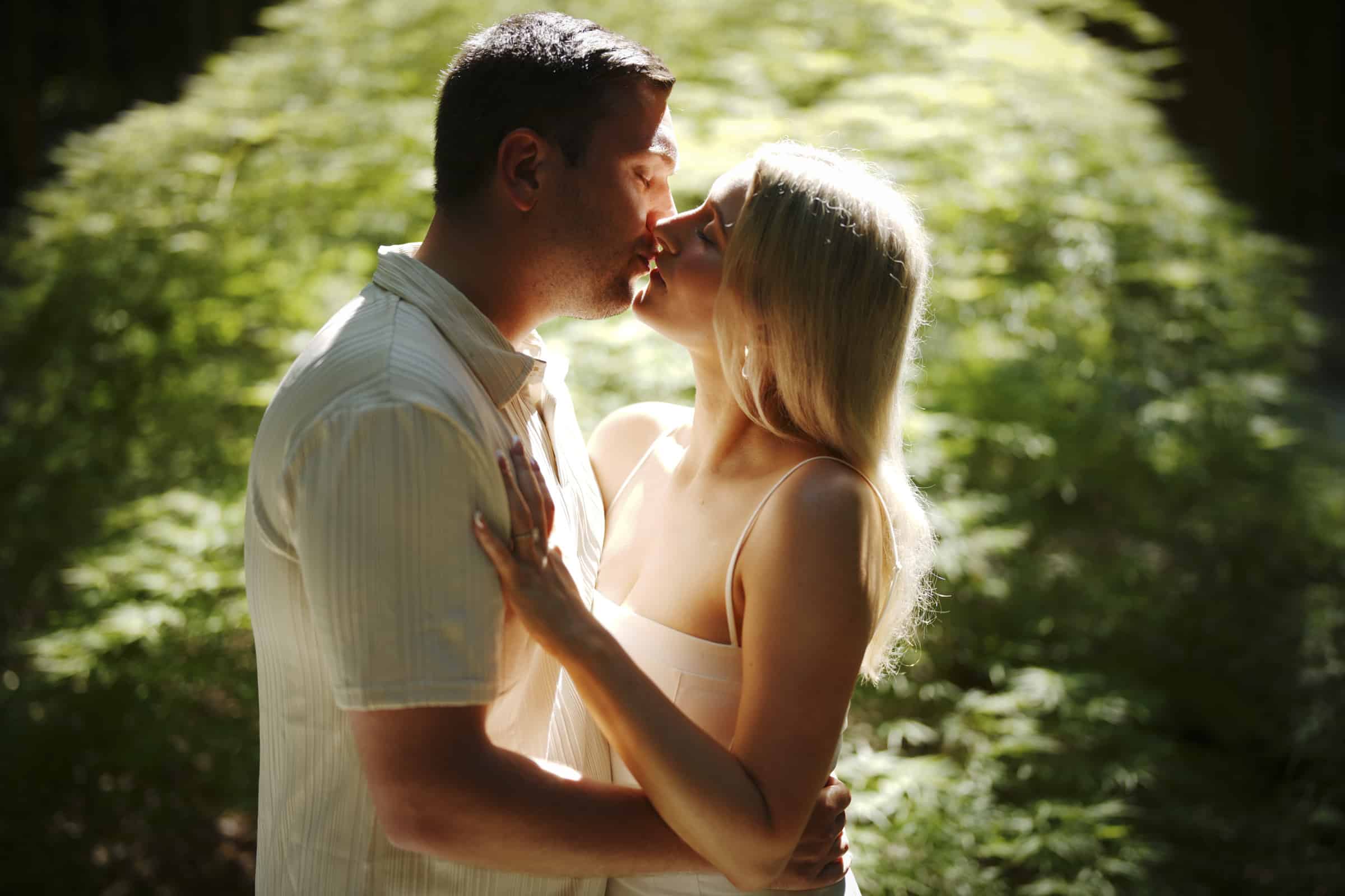 A man and woman gently kiss while standing close together in a sunlit, green outdoor setting, surrounded by lush foliage.