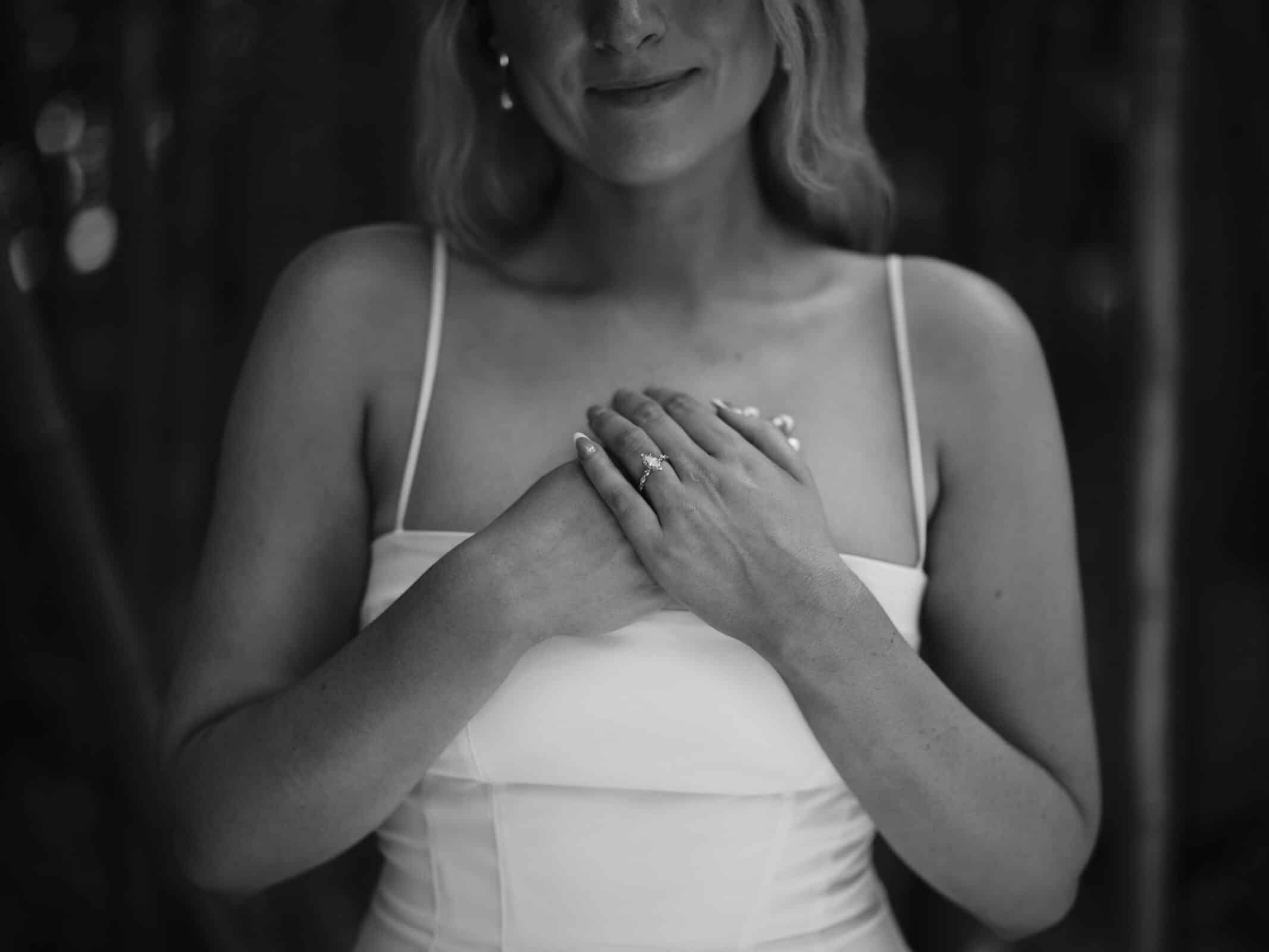 A woman in a white dress stands outdoors with her hands clasped over her chest, displaying a ring on her finger. The image is in black and white, and her face is partially visible, showing a gentle smile.